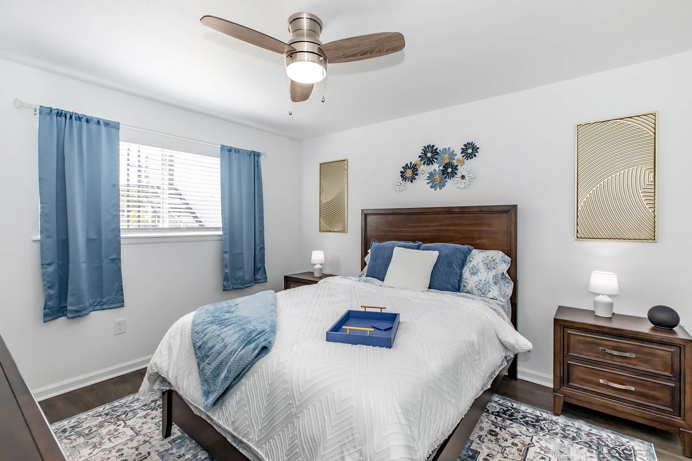 A cozy bedroom featuring a wooden bed with blue and white bedding, two nightstands with lamps, and decorative wall art. The room has light blue curtains on a window, a ceiling fan, and a rug underneath. The overall decor is modern and inviting, creating a serene atmosphere.