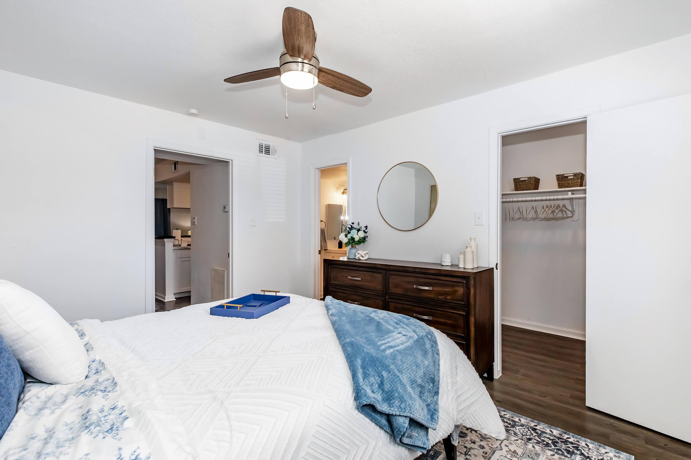 A cozy bedroom featuring a bed with white and blue linens, a wooden dresser, and a round mirror on the wall. A ceiling fan is overhead, and there are white doors leading to a closet and another room in the background. Decorative items and a blue throw blanket add a welcoming touch.
