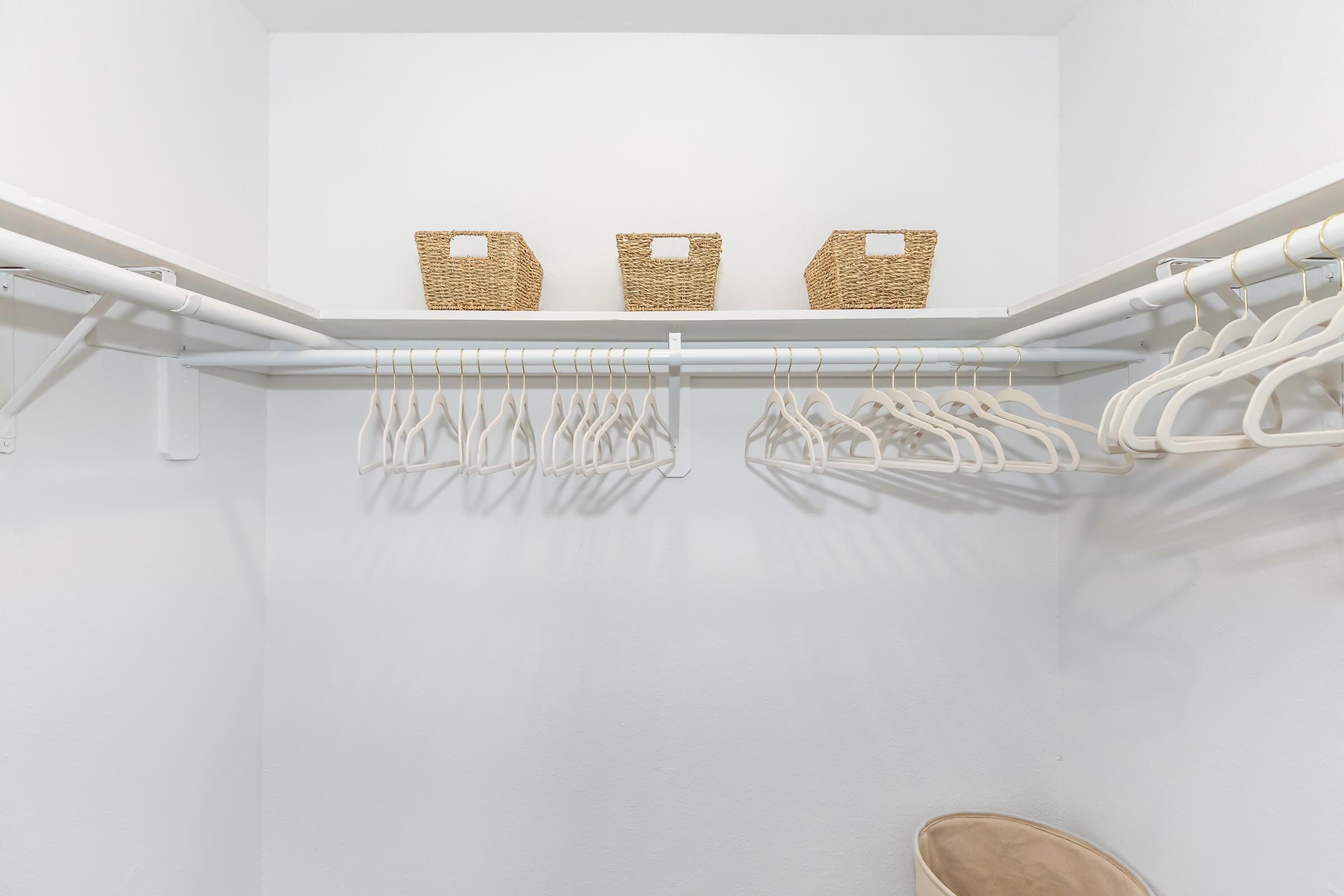 A clean, organized closet featuring a white interior. Two shelves hold three wicker storage baskets, while below, multiple white hangers are neatly arranged on a rod. There is a light, round beige container on the floor, contributing to the tidy atmosphere.