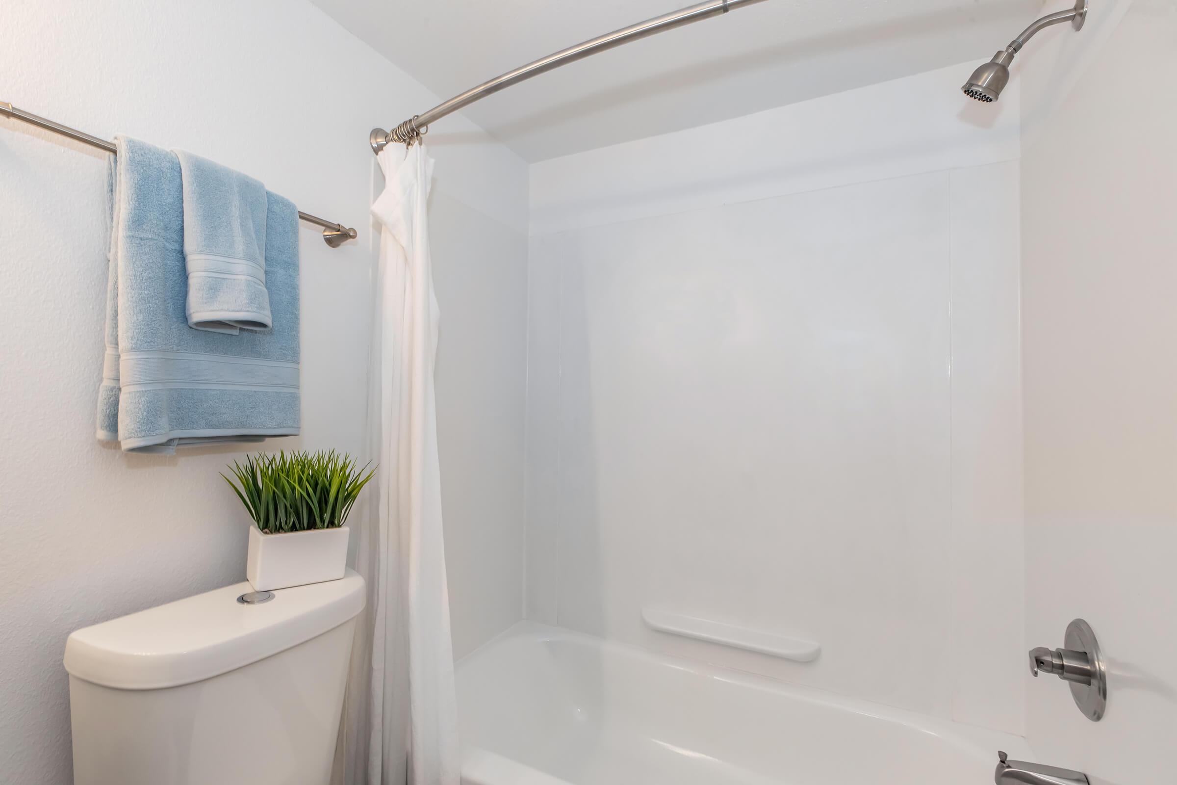 A clean bathroom featuring a bathtub with a white shower curtain, a blue towel hung on a rack, and a potted plant on the edge of the bathtub. The walls are smooth and painted white, creating a bright and fresh atmosphere.
