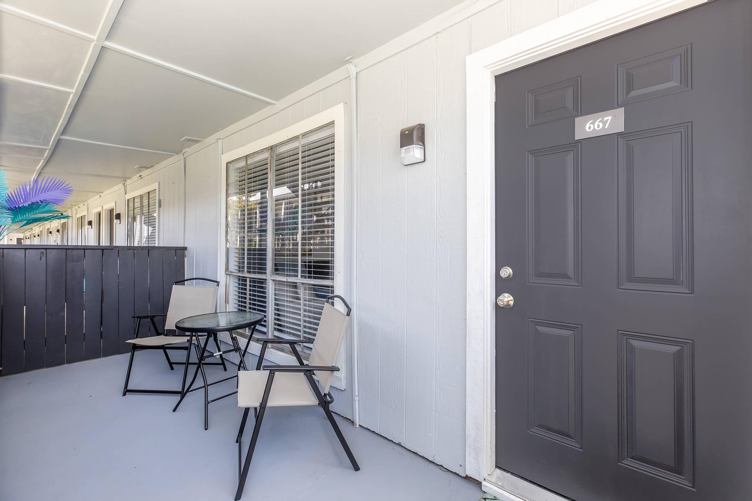 A gray exterior view of an apartment door numbered 667, with a small seating area featuring two chairs and a round table. The door is flanked by windows with blinds, and the entrance is covered by a awning or overhang.