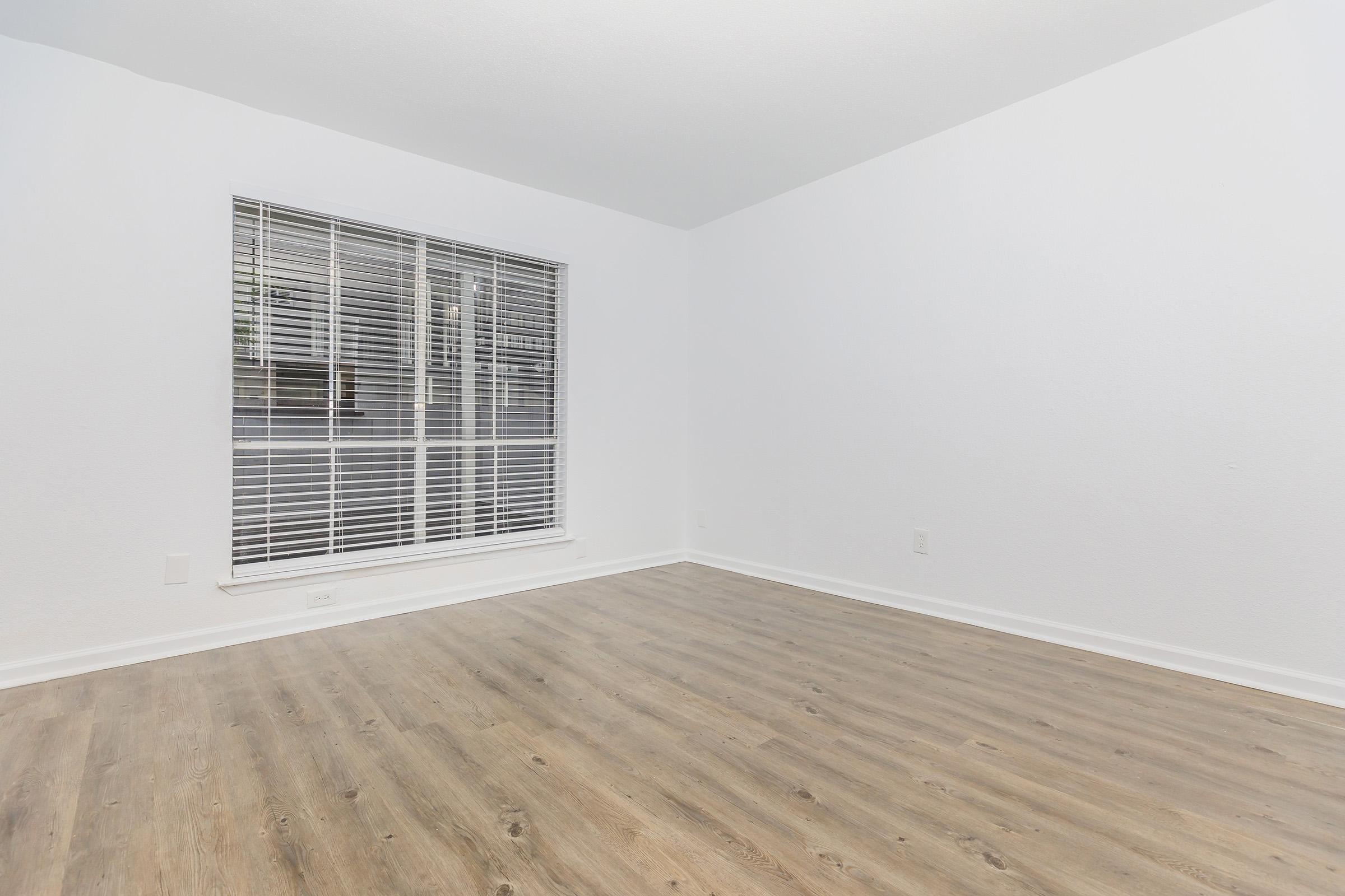 A spacious, empty room with white walls and a large window featuring vertical blinds. The floor is covered in light-colored wood laminate. The space appears bright and clean, ideal for furnishing or decorating.