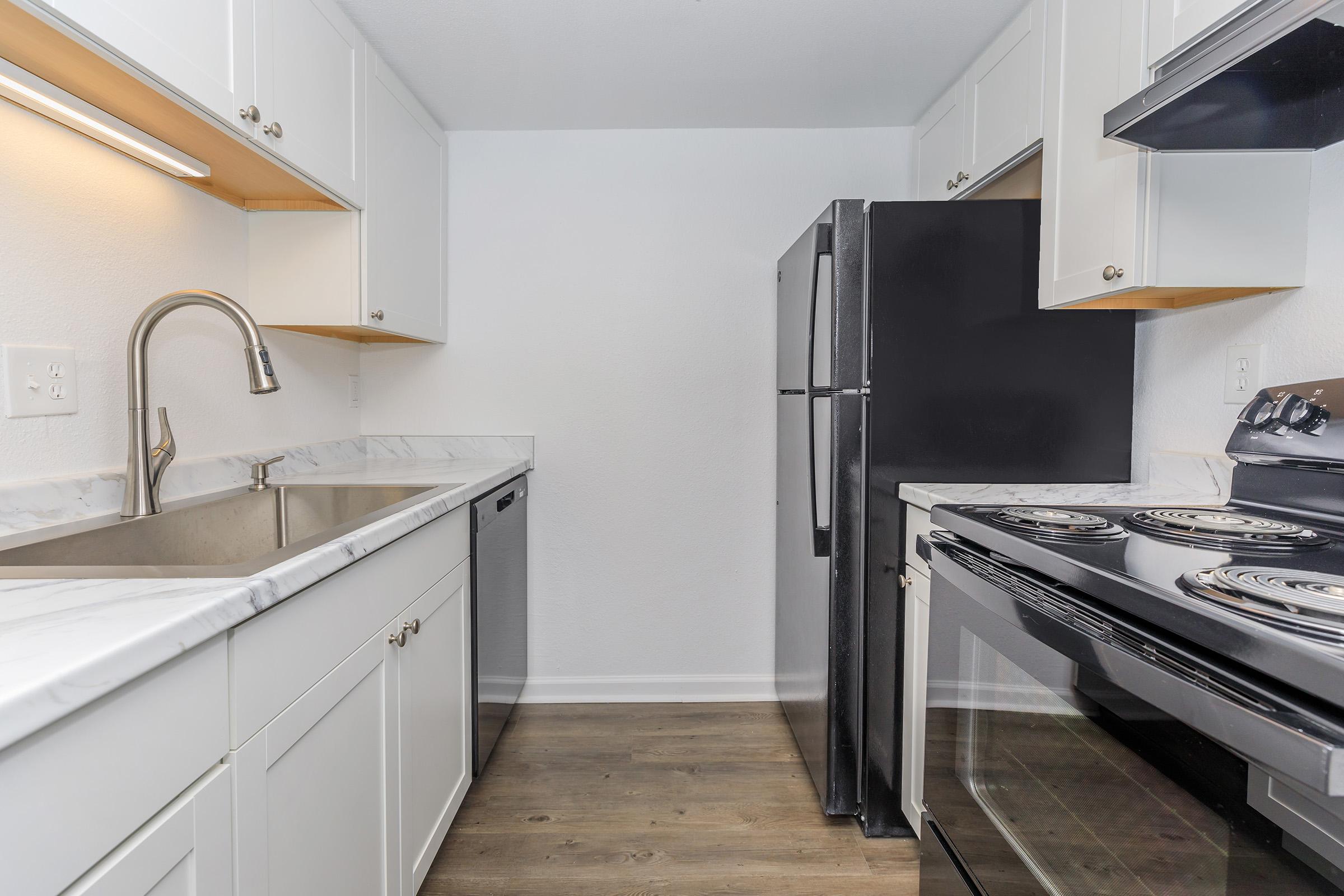 Modern kitchen featuring white cabinetry, a stainless steel sink, and marble countertops. Includes a black refrigerator, a dishwasher, and a black stove with four burners. The floor is a light wood finish, and the walls are painted white, creating a bright and clean space.