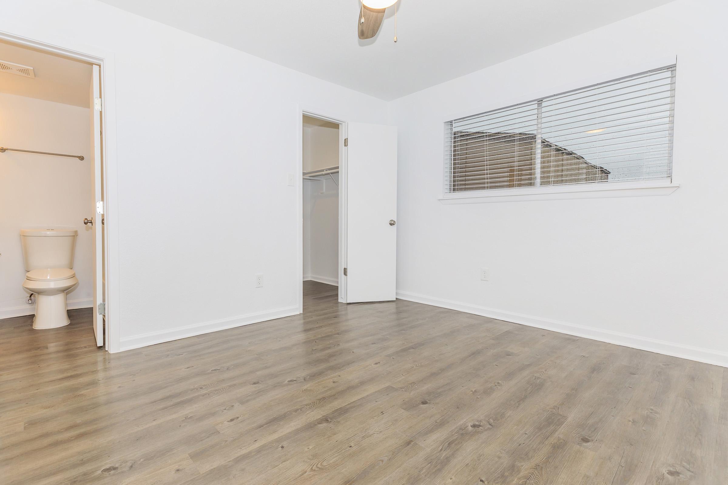 Empty room with white walls and a light-colored wooden floor. A closed door on the left leads to a bathroom, which has a toilet visible. An open door on the right leads to a closet. A window with blinds allows natural light to enter the space. The ceiling has a ceiling fan installed.