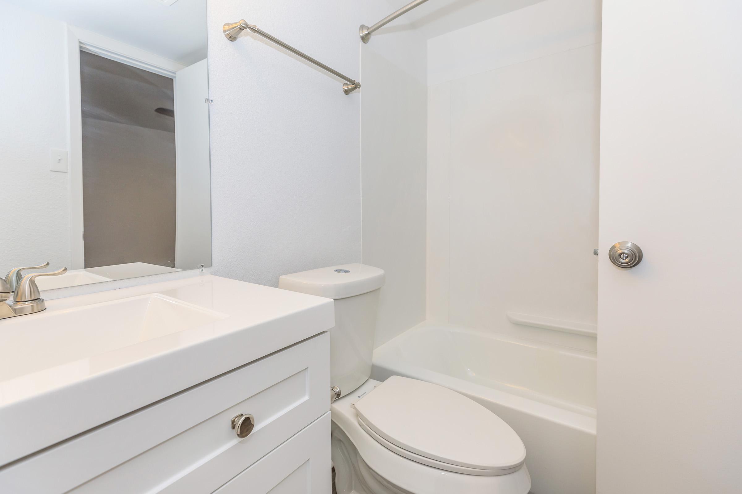 A clean, modern bathroom featuring a white vanity with a sink, a toilet, and a bathtub with a shower. The walls are painted white, creating a bright and open feel. A mirror hangs above the sink, and a door leads to another room in the background.