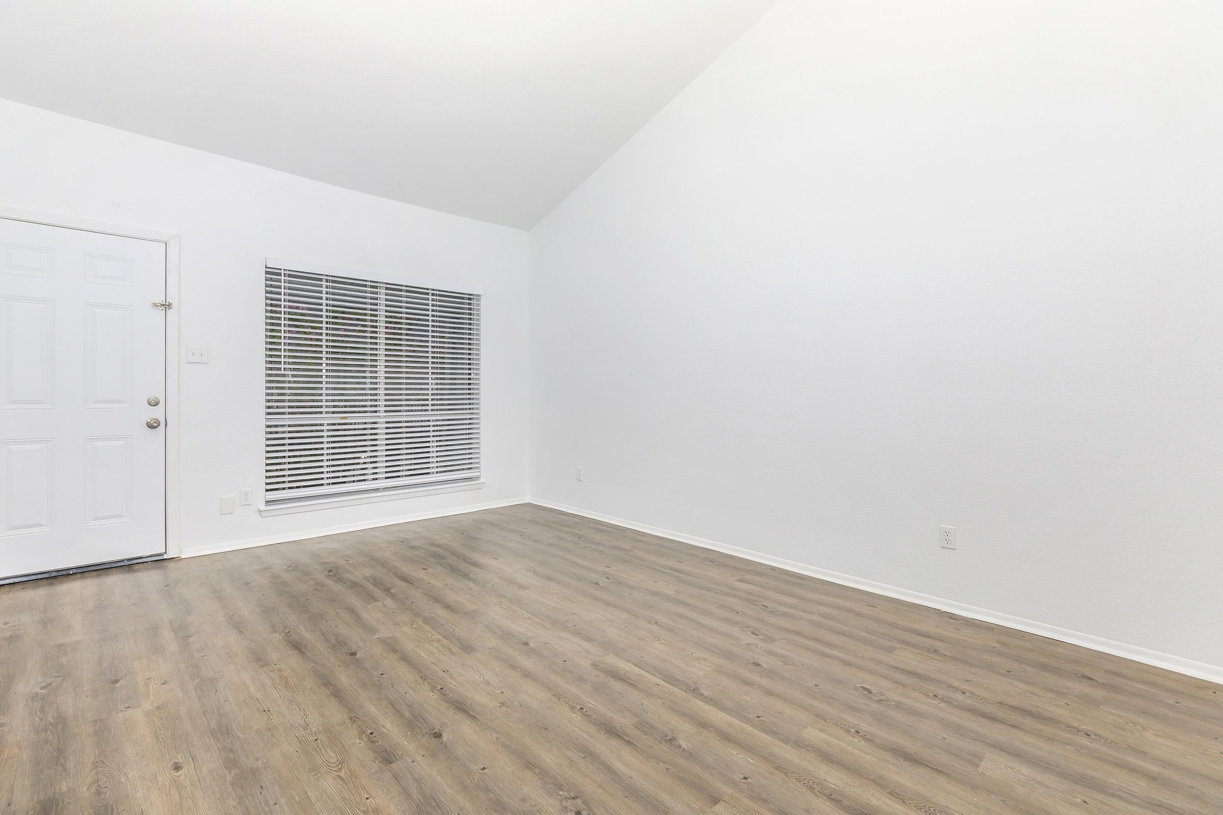 A spacious, well-lit room featuring a high ceiling and wooden flooring. It has a front door on the left and a large window with blinds on the right, allowing natural light to brighten the space. The walls are painted white, creating a clean and open atmosphere.