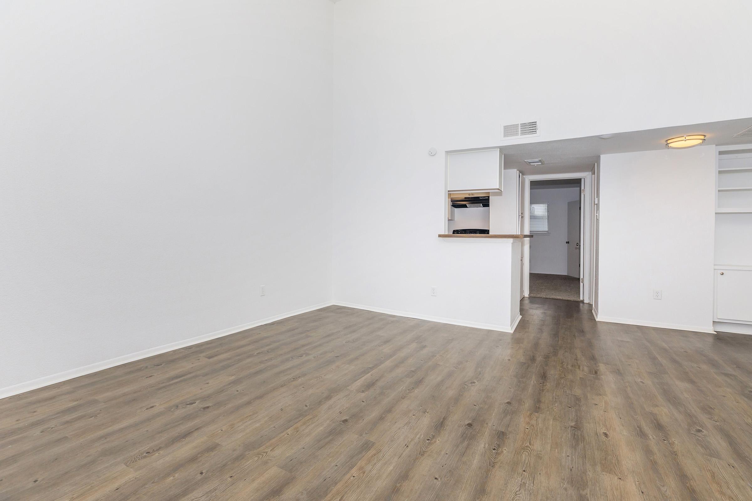 A spacious, minimalist living area with light-colored walls and wooden flooring. The room features a high ceiling, an open layout with a kitchen area visible in the background, and a doorway leading to another room. Natural light fills the space, creating a bright and airy atmosphere.