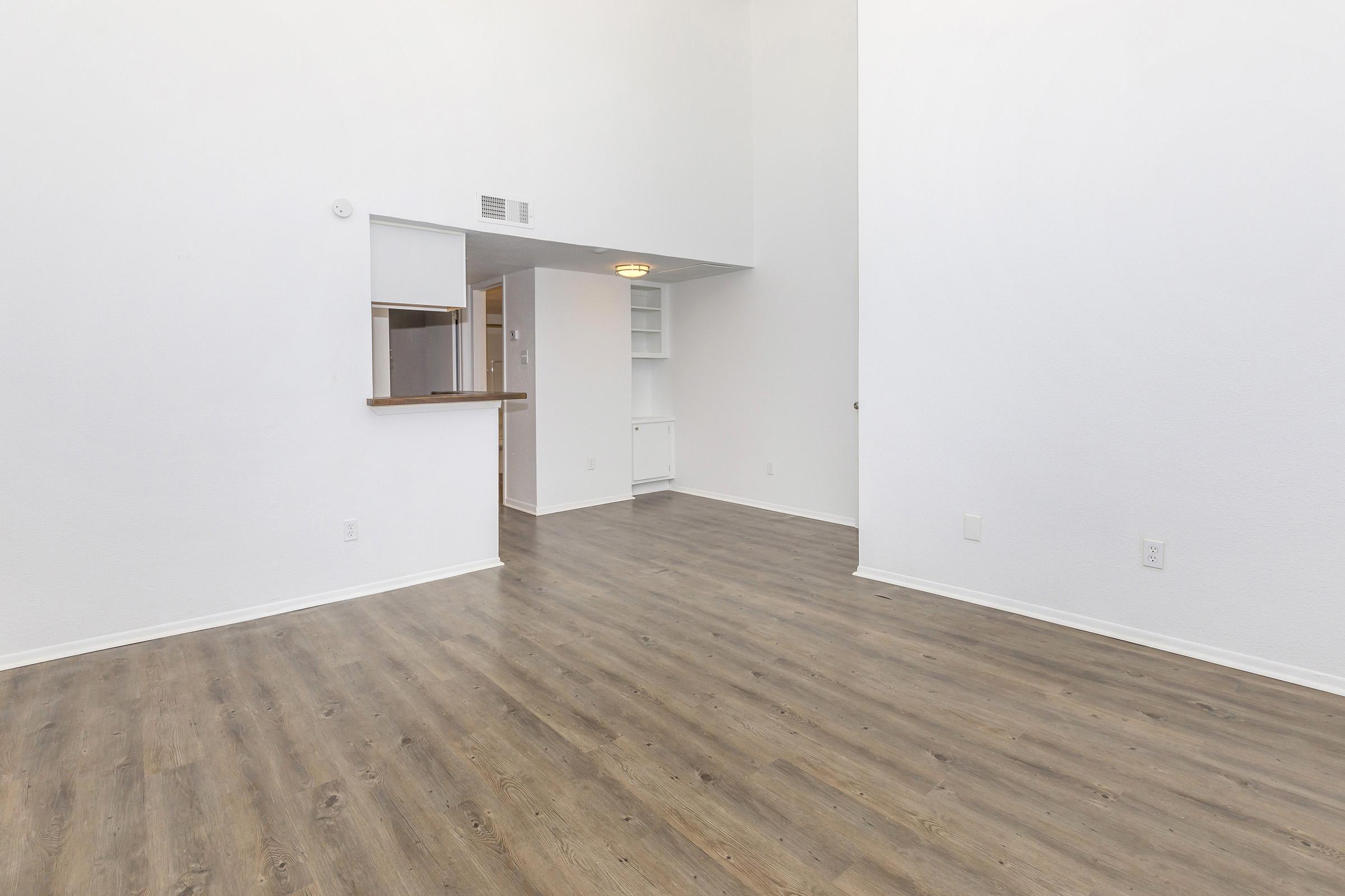 A spacious, empty room with light-colored walls and a high ceiling. The floor is made of wood or laminate, and there is a small bar area that opens to a kitchen. A built-in shelf is visible on the wall, and natural light fills the space, giving it a bright and airy feel.