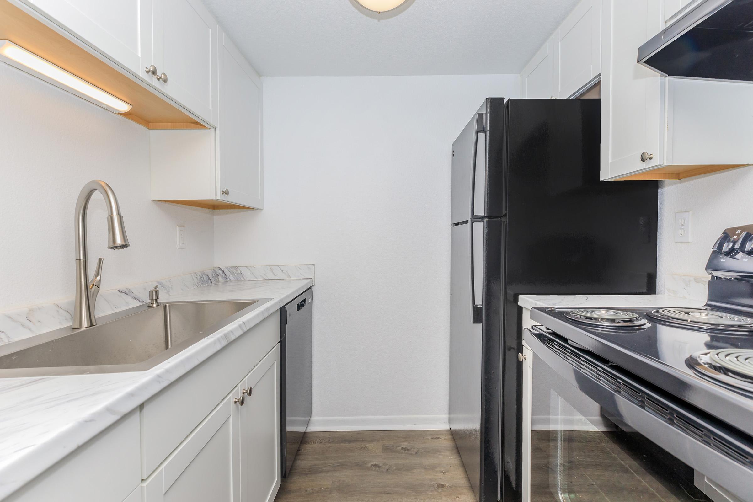 Modern kitchen featuring white cabinetry, a stainless steel sink with a sleek faucet, black refrigerator, and black stove. The countertops are made of marble, and the space has a clean, minimalist design with ample lighting and a tidy appearance.