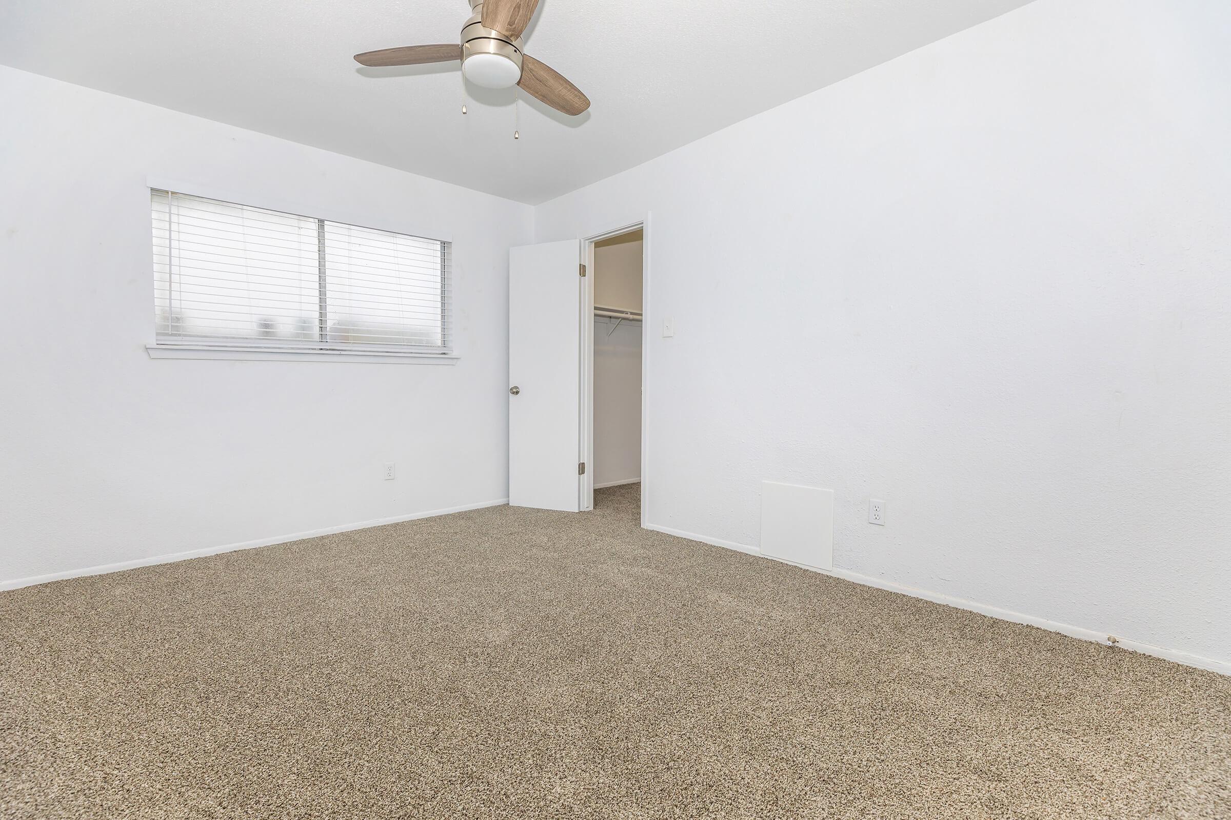A bright, empty room featuring beige carpet flooring, white walls, and a ceiling fan. A window with horizontal blinds allows natural light, and there is an open door leading to a small closet. The minimalistic design and neutral colors give a sense of spaciousness and calm.