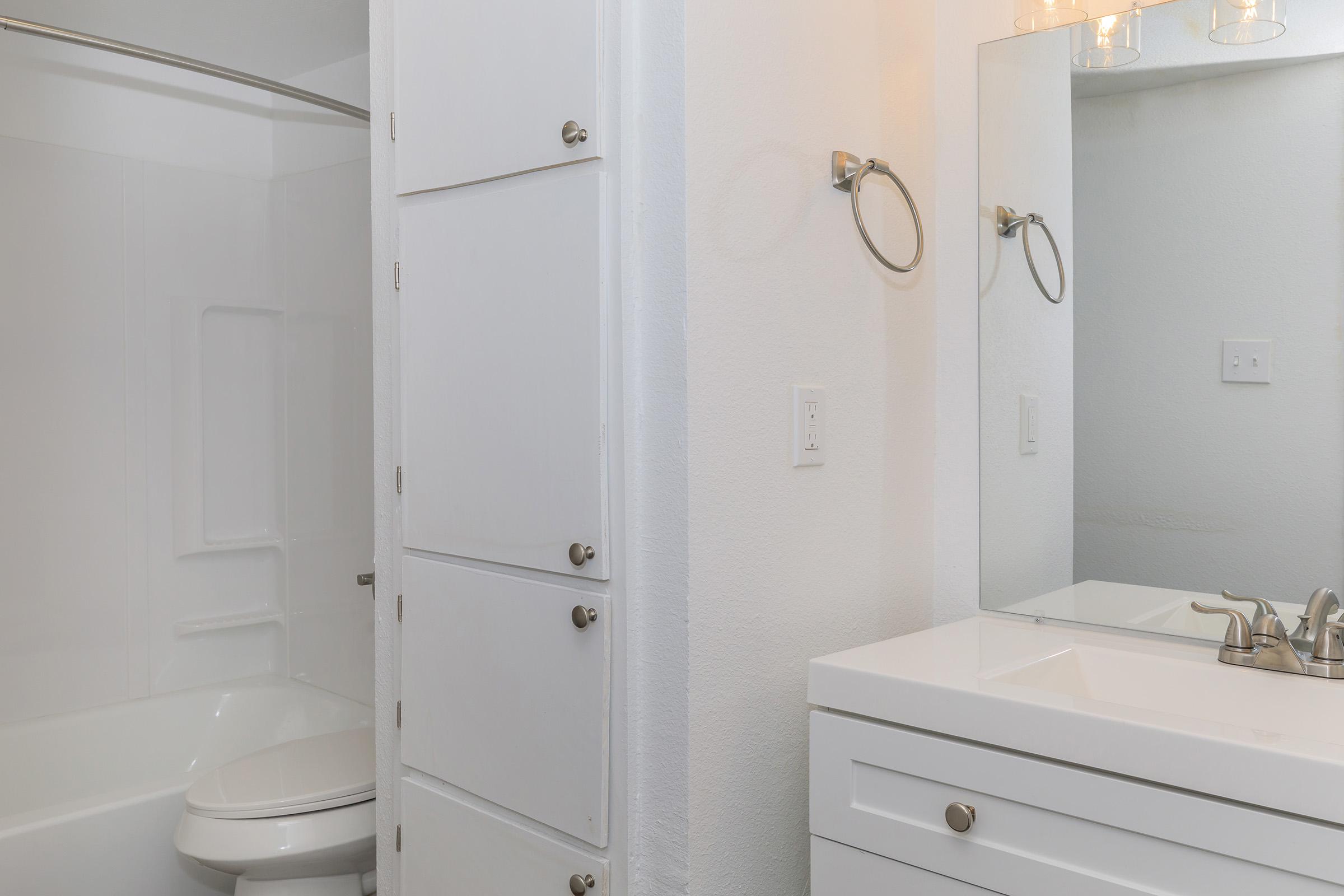 A modern bathroom featuring a white bathtub with a shower, a sink with a mirror above, and a cabinet. The walls are painted white, and the overall décor is clean and contemporary, with silver fixtures and a minimalist design.