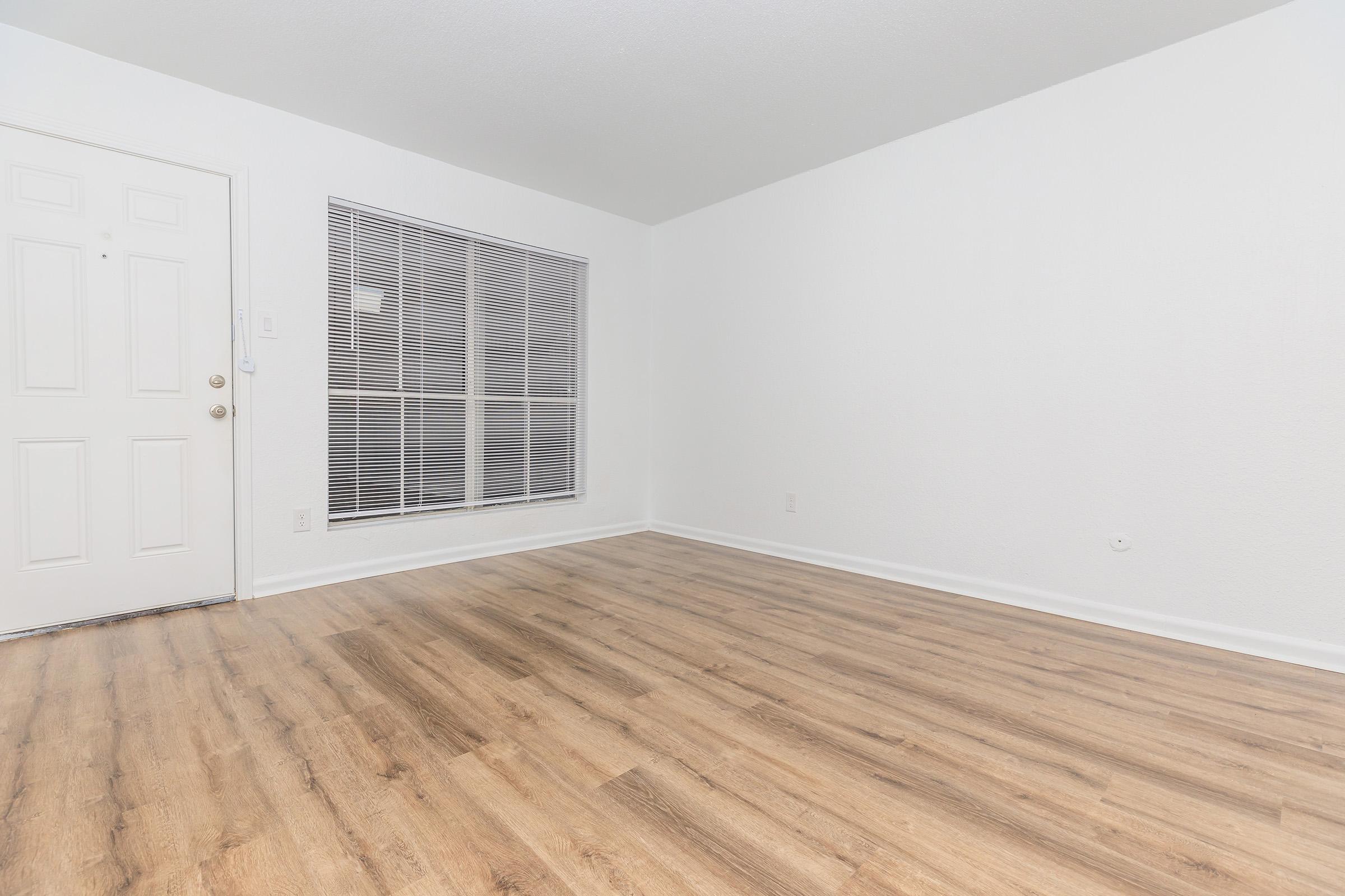 A spacious, empty room with white walls and a light wood-like floor. A front door is visible on the left, and a large window with blinds lets in natural light. The room has a minimalist feel, ideal for various interior design possibilities.