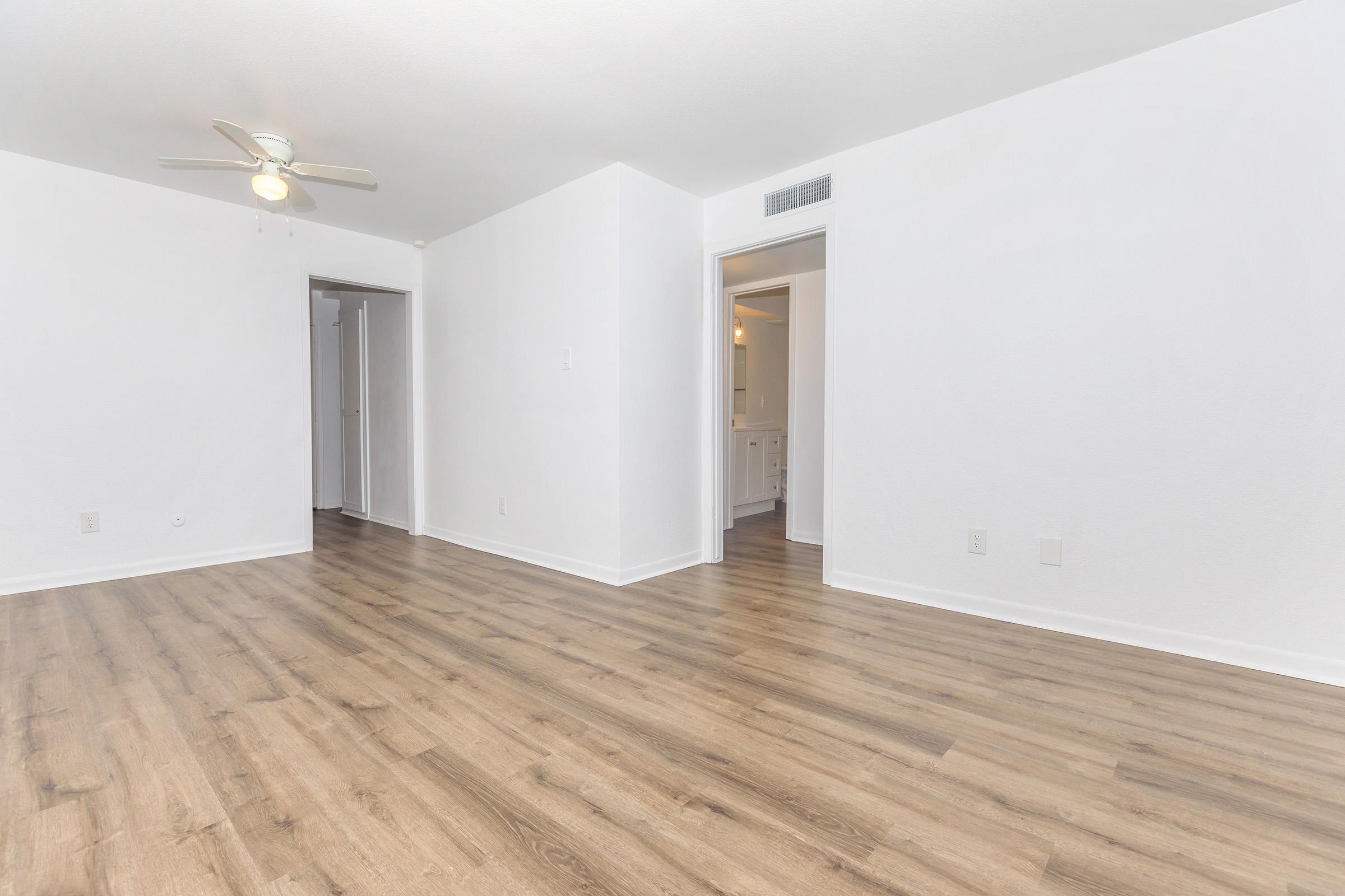 A spacious, empty room with light-colored walls and wooden flooring. A ceiling fan is visible, and there are doorways leading to adjacent areas. The room has a bright and clean appearance, suitable for various uses or furnishings.