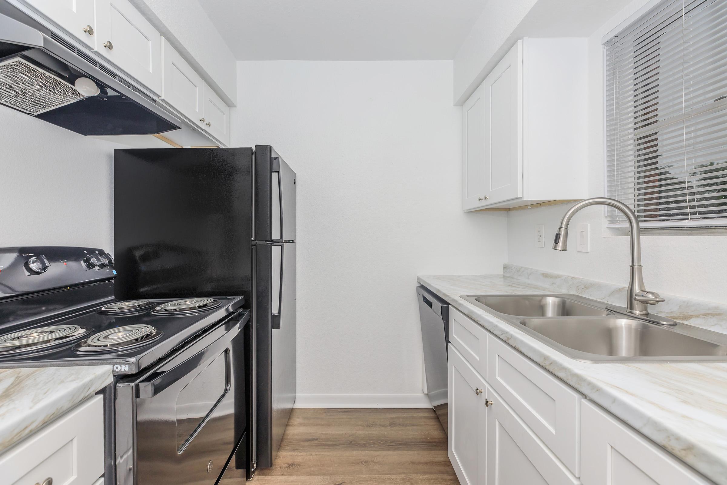 A modern kitchen featuring white cabinetry, a black refrigerator, a black oven, and a double sink. The countertops are light-colored, and there is a window providing natural light. The flooring is wood-like, creating a cozy atmosphere.