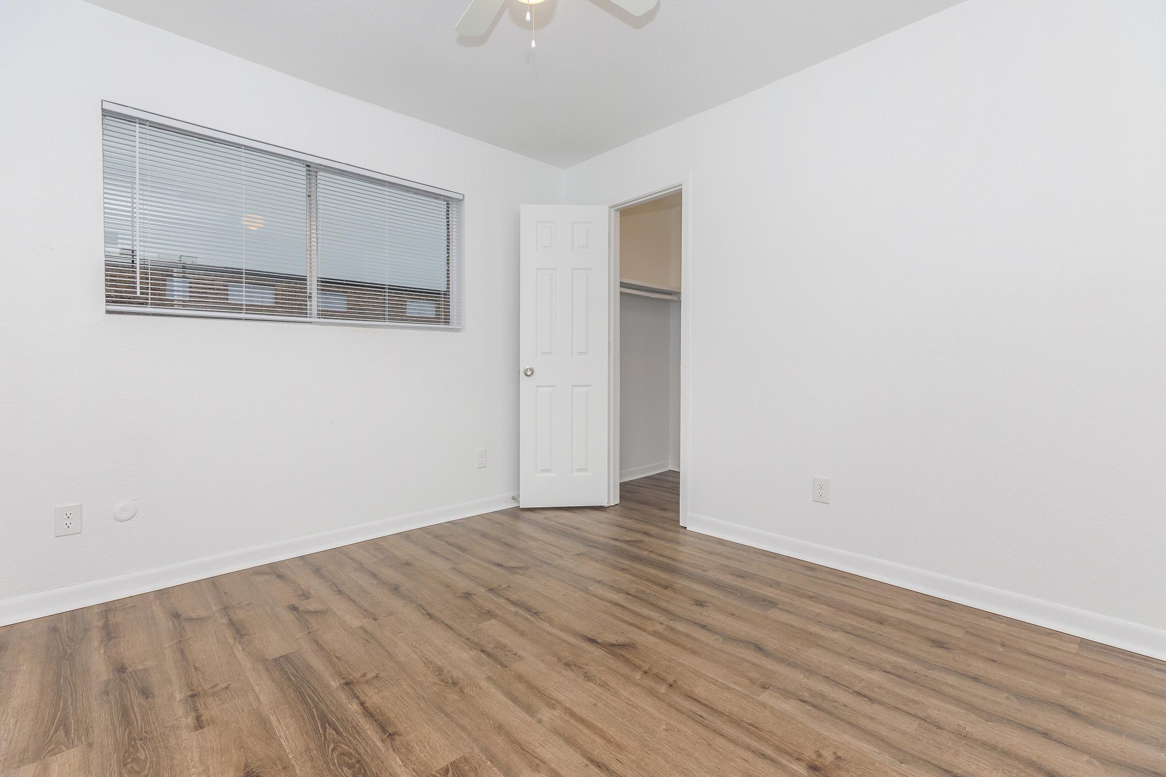 A well-lit room featuring light-colored walls, a ceiling fan, and a large window with blinds. The flooring is made of light wood, and there is a doorway leading to a closet. The overall atmosphere is spacious and clean, suitable for various uses.