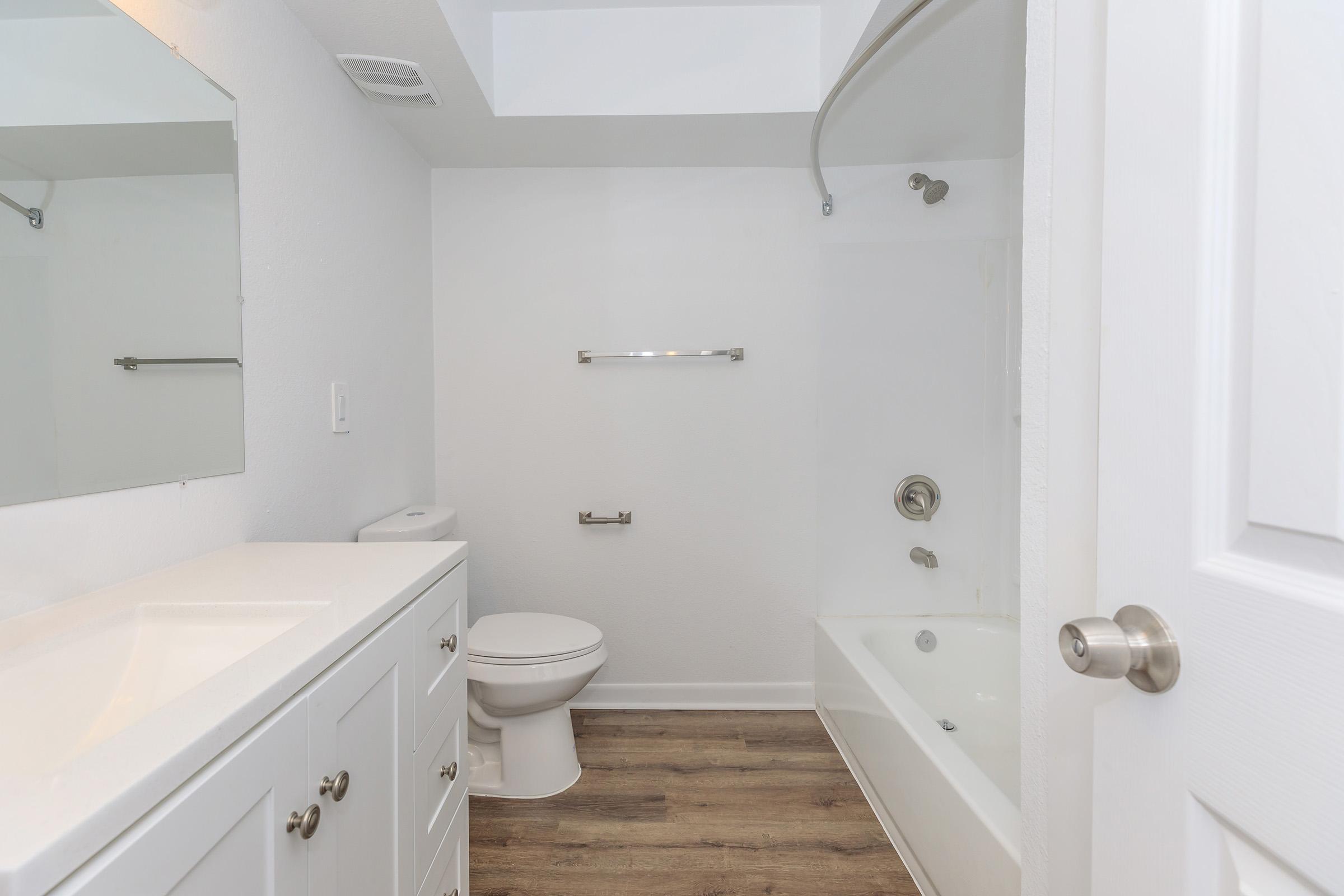 A clean and modern bathroom featuring a white bathtub with a shower, a wall-mounted mirror above a white vanity cabinet, and a toilet. The walls are painted white, and the flooring is a light wood. Natural light enters through a small window on the ceiling, creating a bright and spacious feel.
