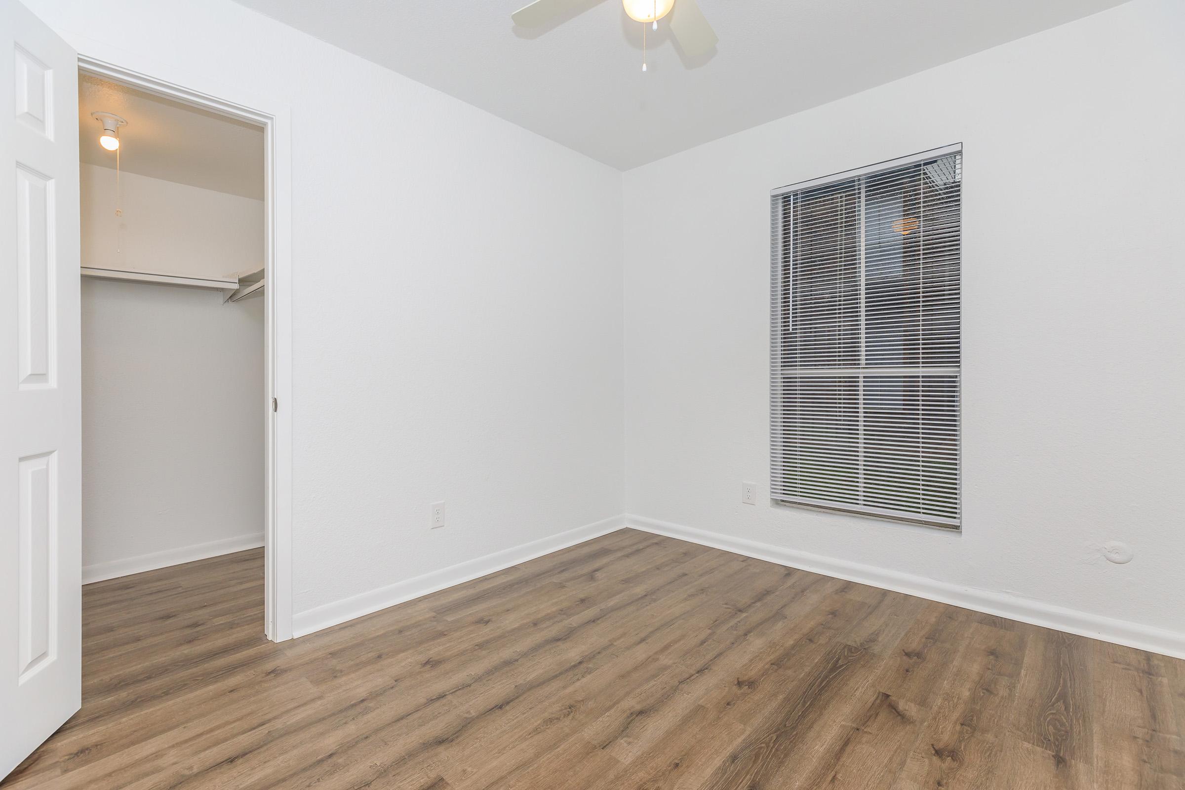 A well-lit, empty room featuring a light-colored wall, a ceiling fan, and a window with blinds. There is a closet to the left, and the floor is covered with laminate wood, giving the space a modern and clean appearance.