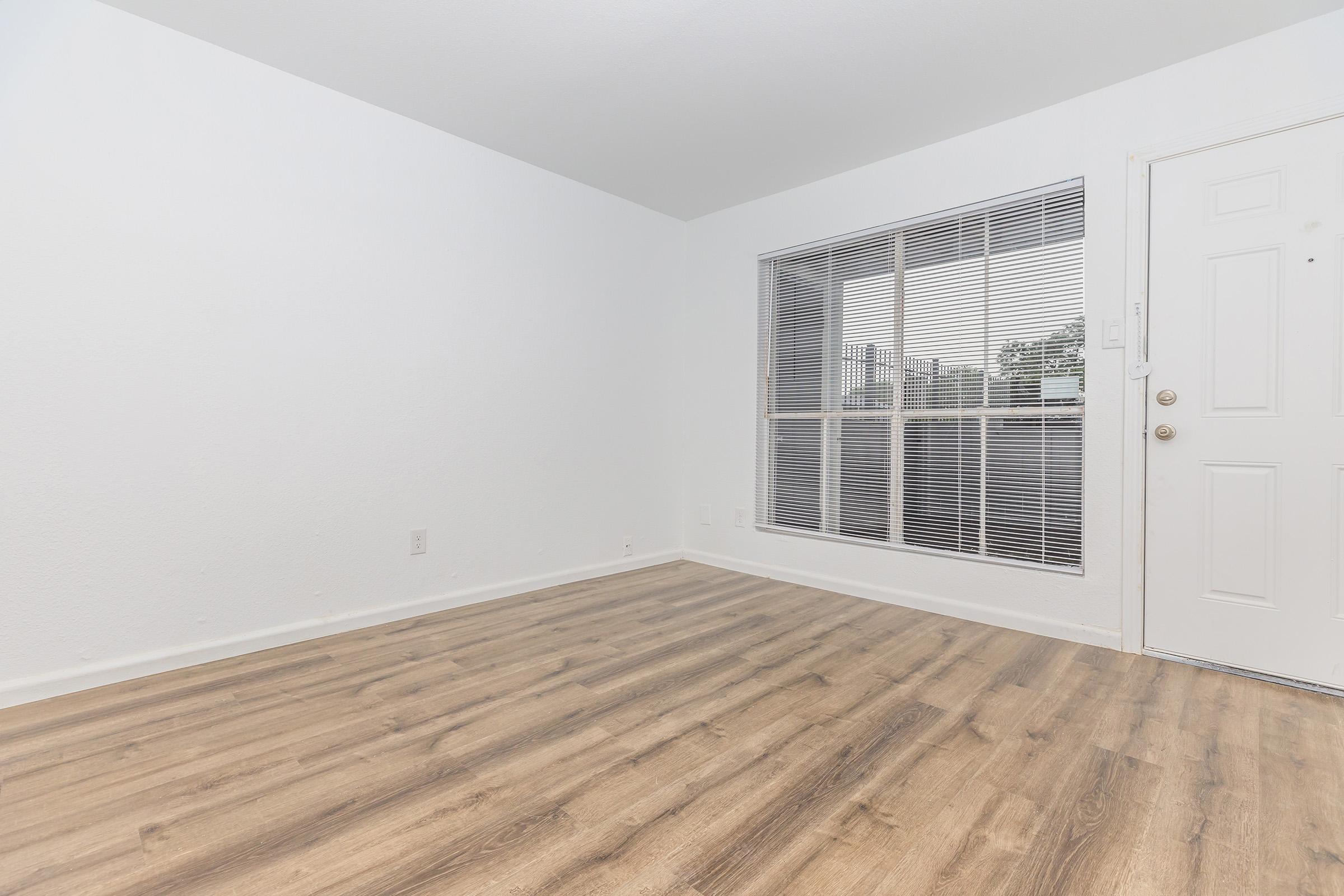 Empty room with light-colored walls and laminate flooring. A large window with blinds lets in natural light, and a front door is visible on the right side. The space appears clean and minimalistic, suitable for various uses such as a living area or office.