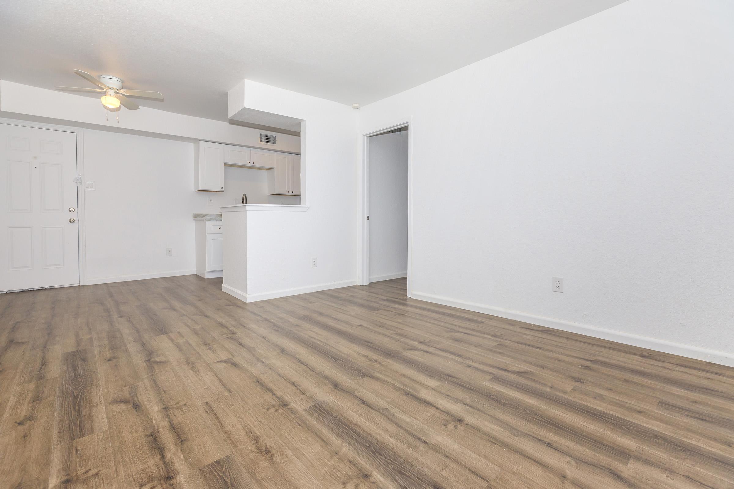 A spacious, empty living area with light-colored walls and modern wood-like flooring. There is a ceiling fan and a small kitchen area visible in the background. The room is well-lit and unfurnished, providing a blank canvas for decoration. A doorway leads to another room on the right.