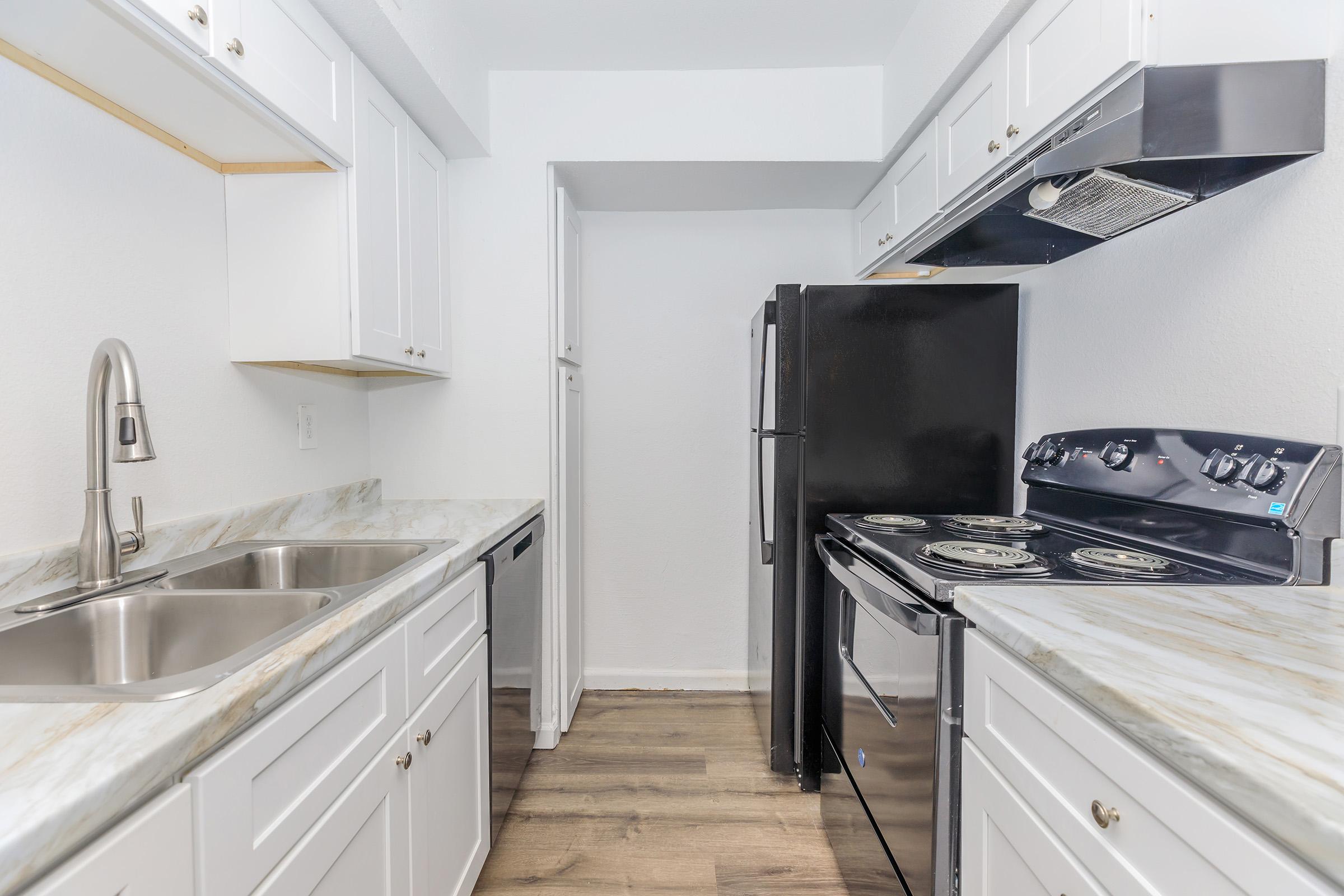 A modern kitchen featuring white cabinetry, a double sink, and a black refrigerator. The countertop has a marble finish, and there is a black stove and oven. The flooring is wood-like, creating a bright and clean atmosphere.