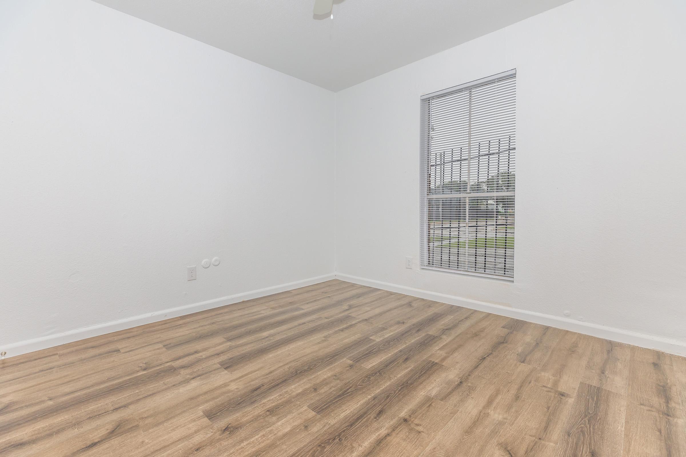Empty room featuring light-colored walls, a wooden floor, and a window with horizontal blinds. Natural light enters through the window, illuminating the space. The room has no furniture or decorations, creating a minimalist appearance.
