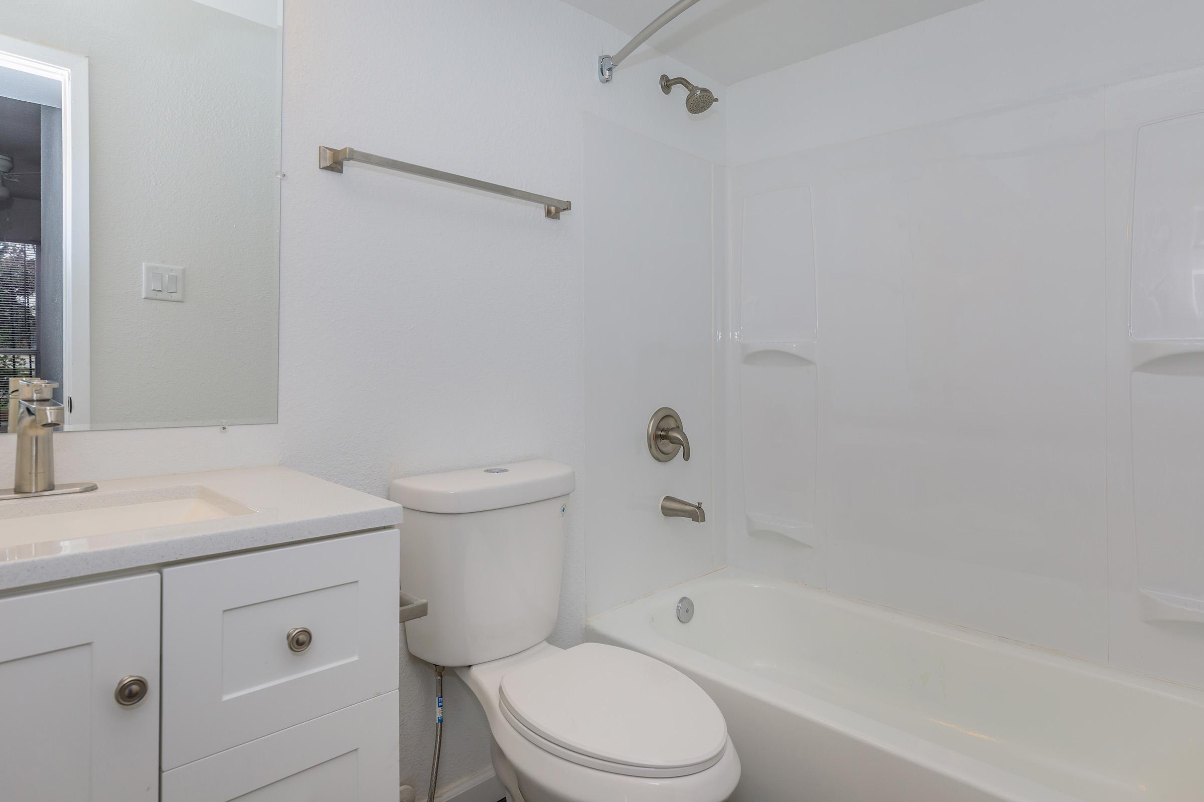A clean and modern bathroom featuring a white bathtub, a shower with a glass door, a toilet, and a sink with a vanity. The walls are painted white, and there is a large mirror above the sink. The overall design is simple and bright, creating a spacious and inviting atmosphere.