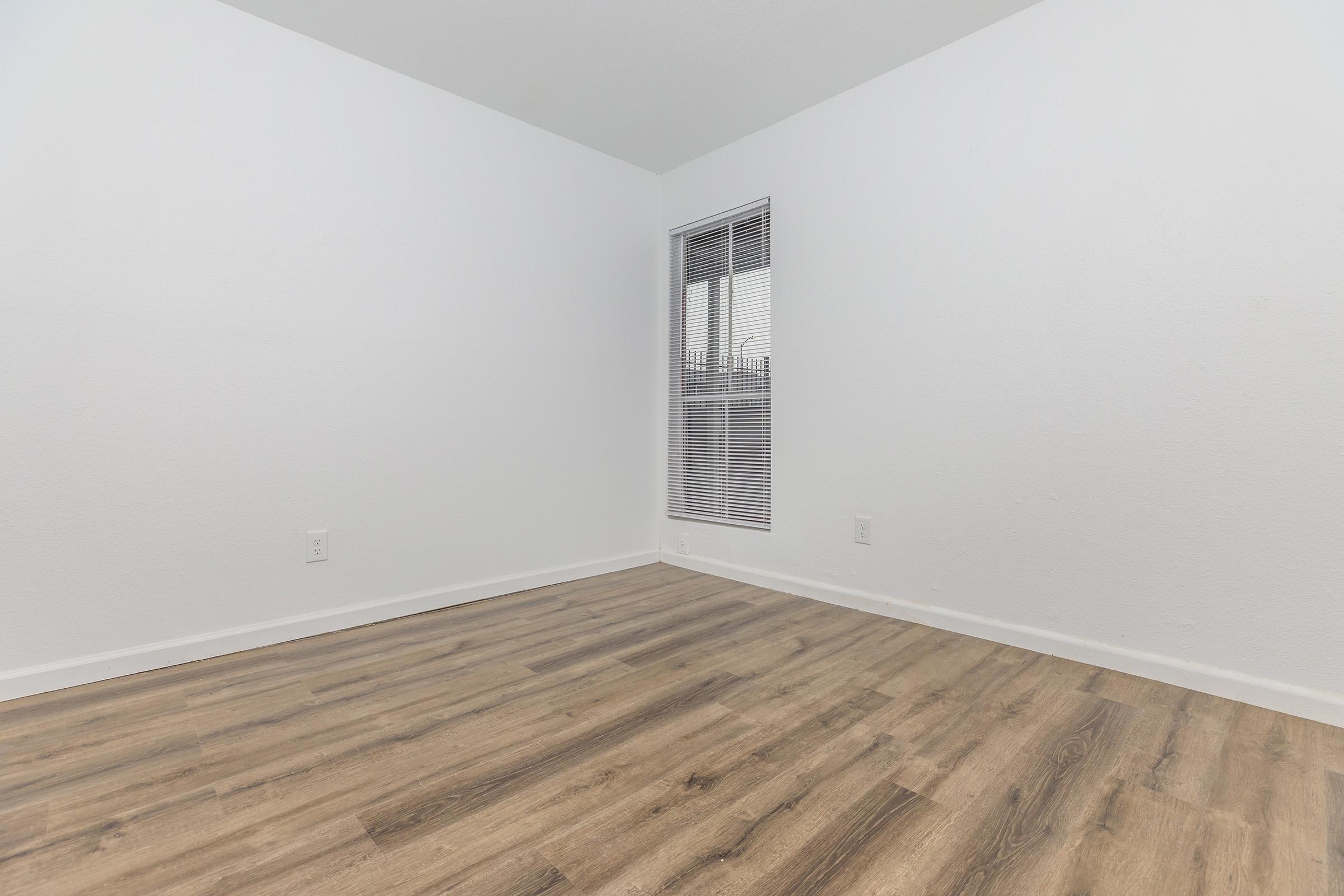 Empty room with white walls and laminate flooring. One window with blinds allows natural light to enter. The space is clean and minimalistic, suitable for various uses or decoration.