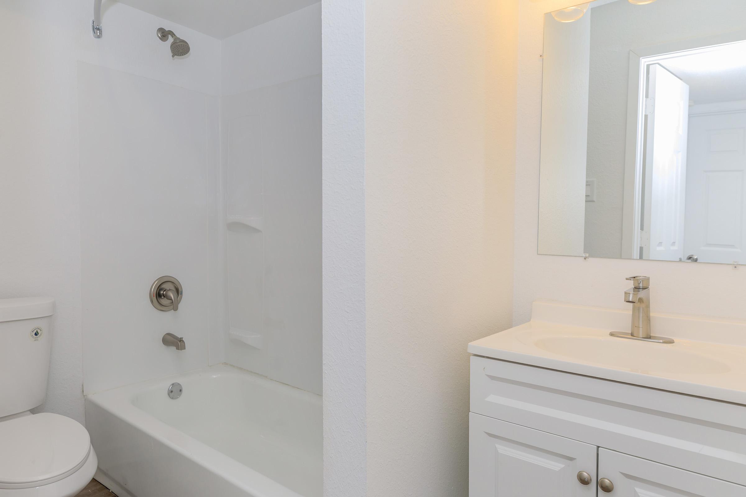 A clean bathroom featuring a white tub and shower, a modern vanity with a sink, a large mirror, and a toilet. The walls are painted in light colors, contributing to a bright and airy atmosphere. The flooring appears to be a light wood-like material, enhancing the overall aesthetic.