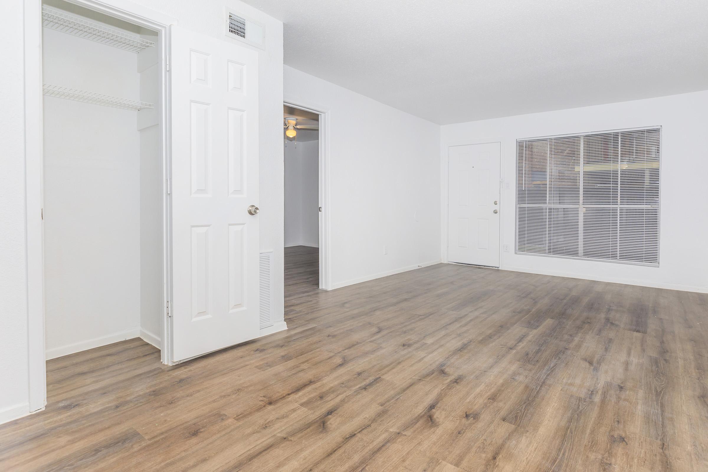 Spacious interior of a well-lit room featuring hardwood floors, white walls, a closet with double doors, a doorway leading to another room, a front door, and a large window with blinds. The layout creates an open and inviting atmosphere.