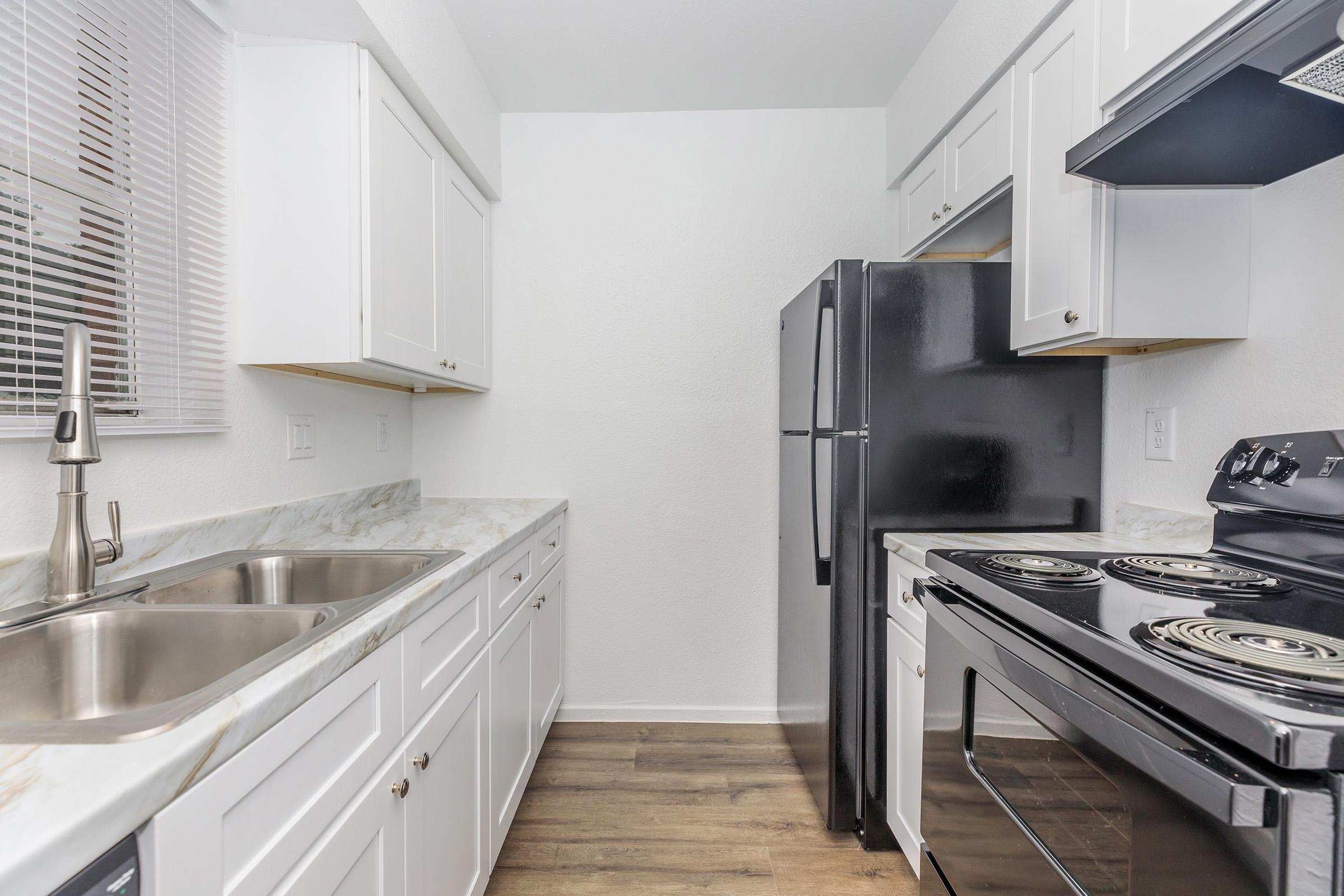 A modern kitchen featuring white cabinets, a black refrigerator, a black stove, and a dual-basin stainless steel sink. The countertops have a light marble design, and the floor is a warm wood tone. Natural light enters through a window with horizontal blinds.