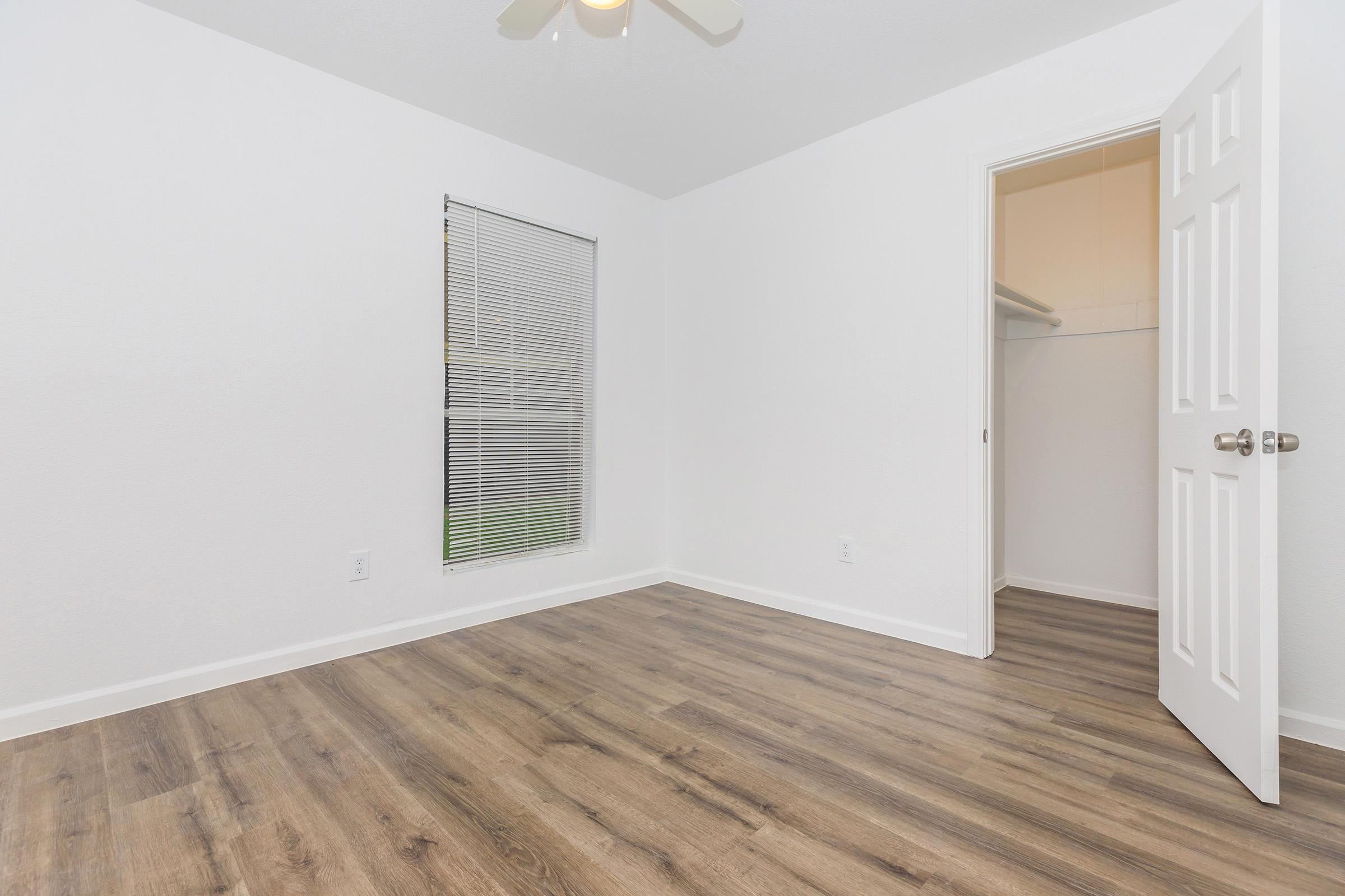 Empty, well-lit room featuring light-colored walls, a single window with blinds, and a closet door slightly open. The floor is covered in light wood laminate. A ceiling fan is visible, adding to the room's airy feel. The space is minimalistic and ready for furnishing.