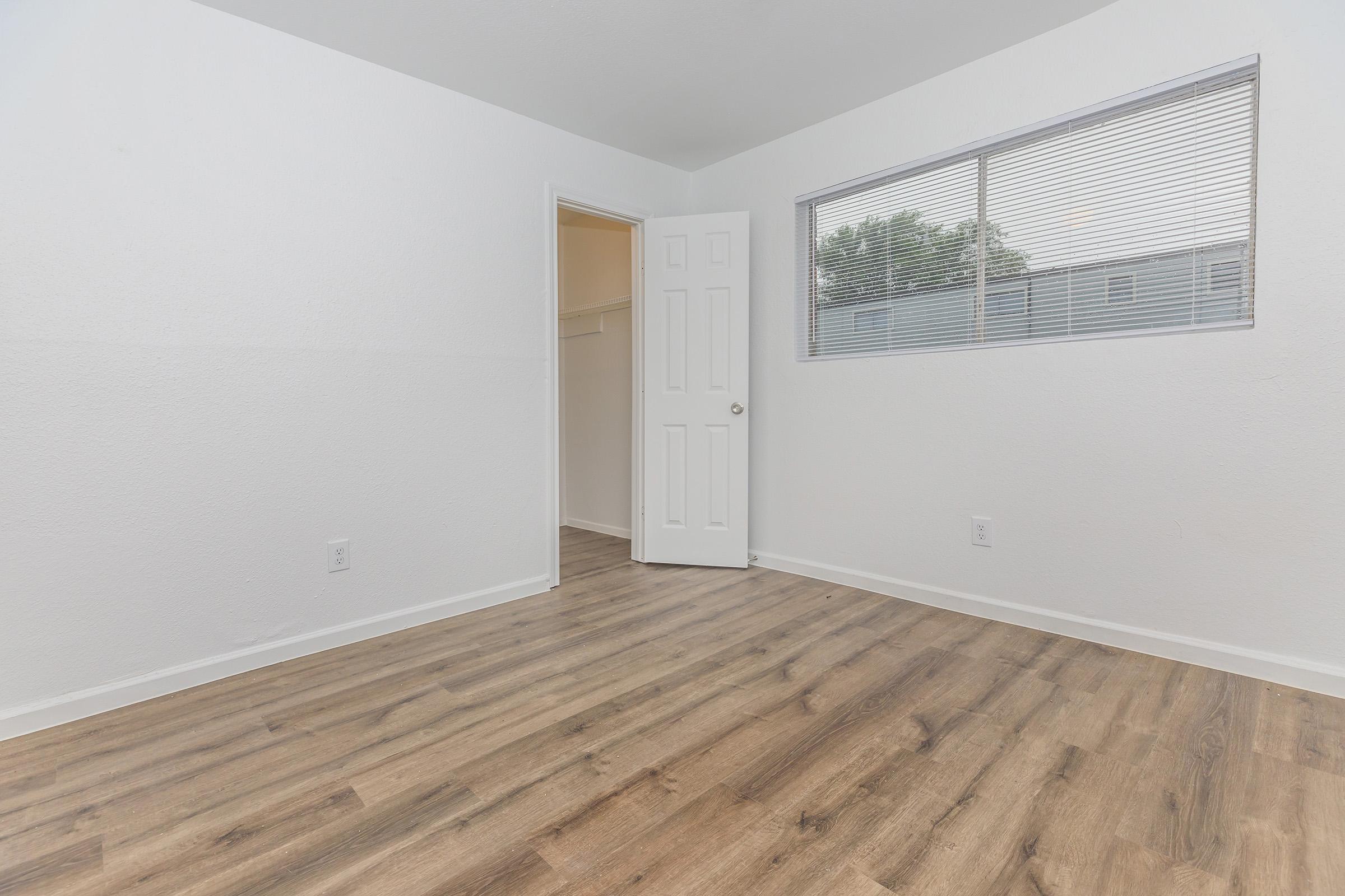 A spacious, empty room with white walls and a large window letting in natural light. The floor features light-colored laminate wood. There is a door on the left leading to another area, and a small closet opening is visible in the corner. The overall ambiance is bright and minimalist.