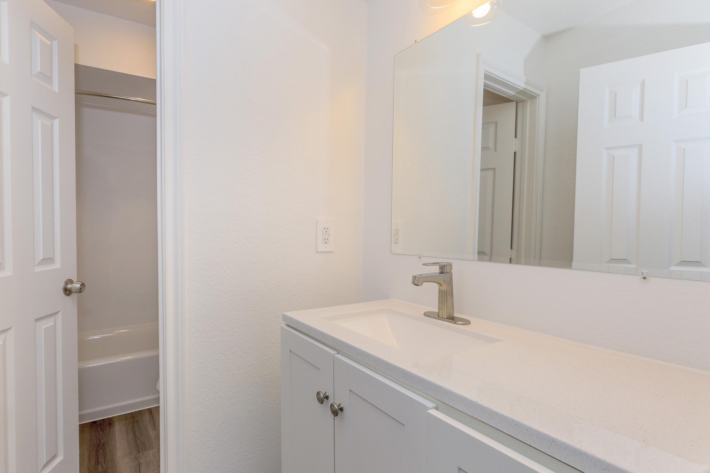A modern bathroom featuring a sleek white vanity with a sink, a large mirror above it, and a door leading to a separate area with a shower or bathtub. The walls are painted in a neutral tone, and the floor displays wood-like tiles, enhancing the clean and bright appearance of the space.