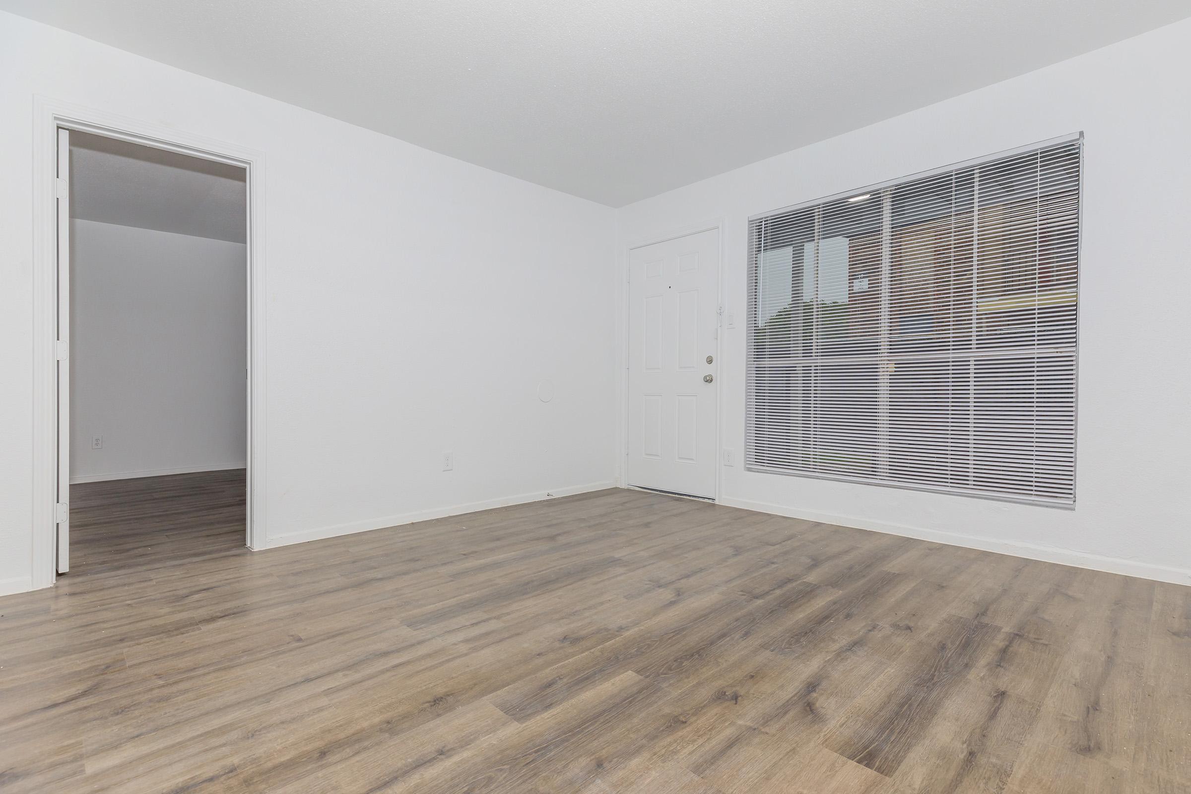 A bright, empty living room featuring light-colored walls and a large window with blinds. The flooring is a light wood-like laminate. There are two doorways, one leading to another room and the other to the entrance. The space is clean and open, ready for furniture and personal decoration.
