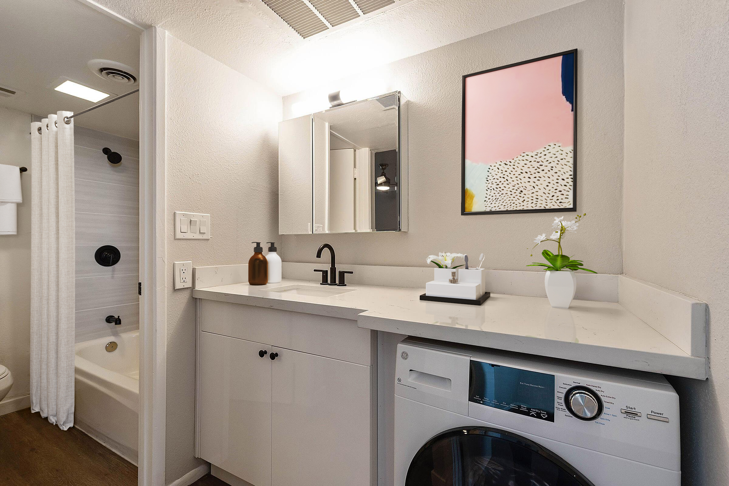 A modern bathroom featuring a double sink vanity with a large mirror, a washing machine, and a small plant. The shower area is visible with a white curtain. The walls are light-colored, and there is an abstract art piece hanging above the sink. The overall design is clean and contemporary.