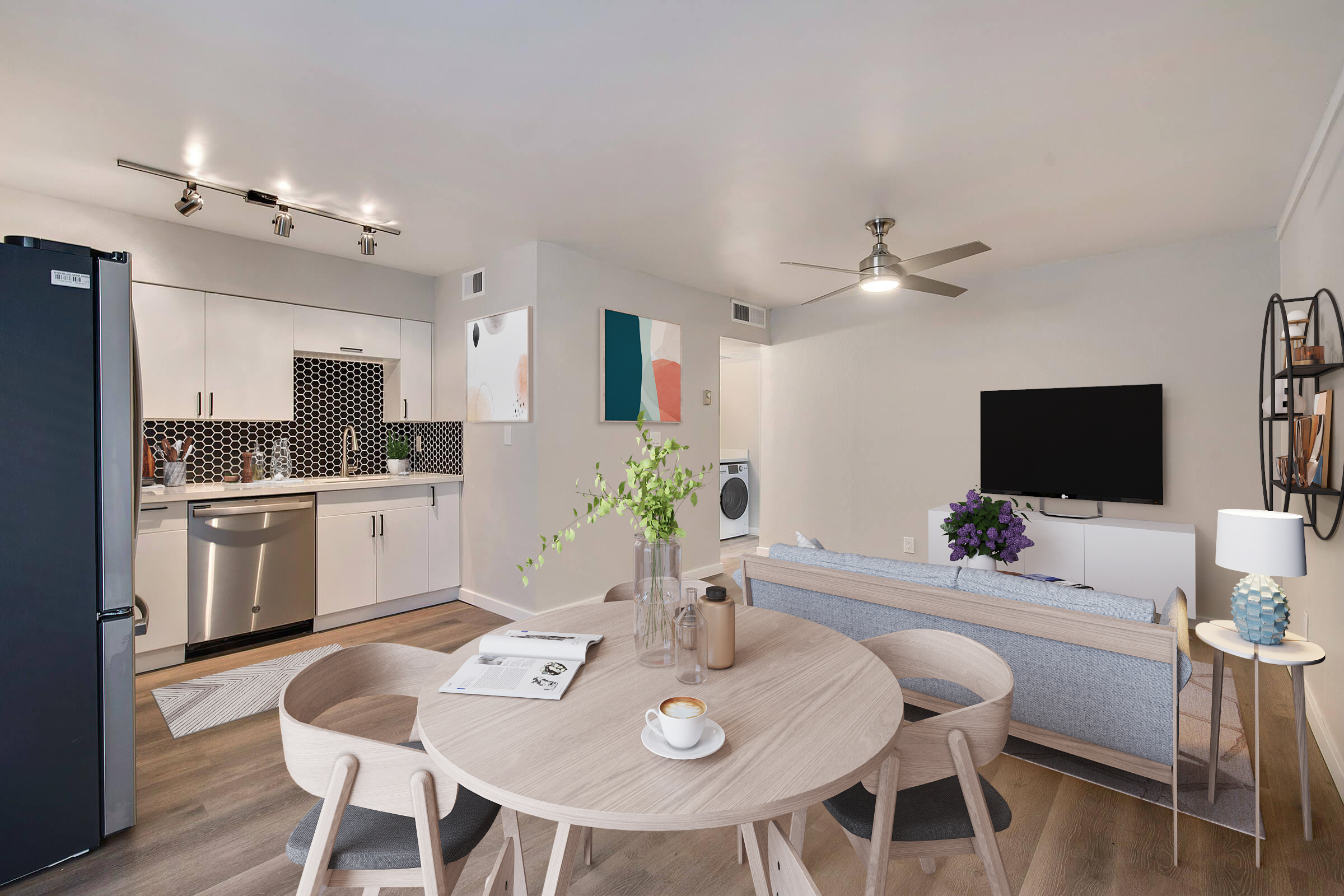 A modern, stylish kitchen and living area featuring a round wooden dining table with chairs, a sleek kitchen with black and white tiles, stainless steel appliances, a sofa, a TV on the wall, and decorative plants. Light-colored walls and warm lighting create an inviting atmosphere.