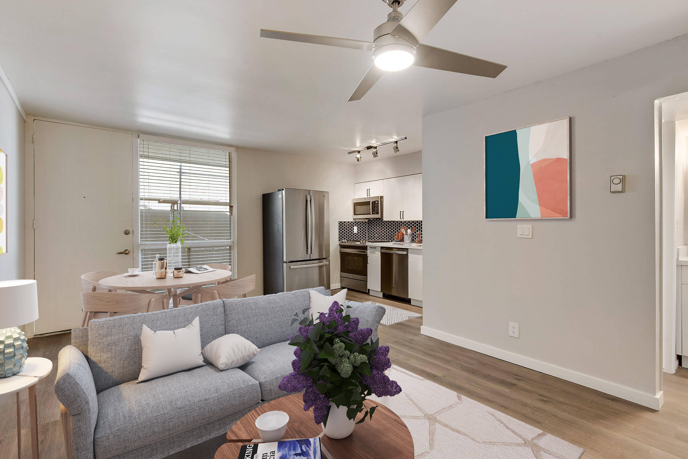 Bright and modern living space featuring a cozy gray sofa adorned with pillows and a vase of purple flowers. A round wooden dining table sits nearby. The kitchen is visible with stainless steel appliances and geometric backsplash. Natural light streams through the window, illuminating the stylish decor.