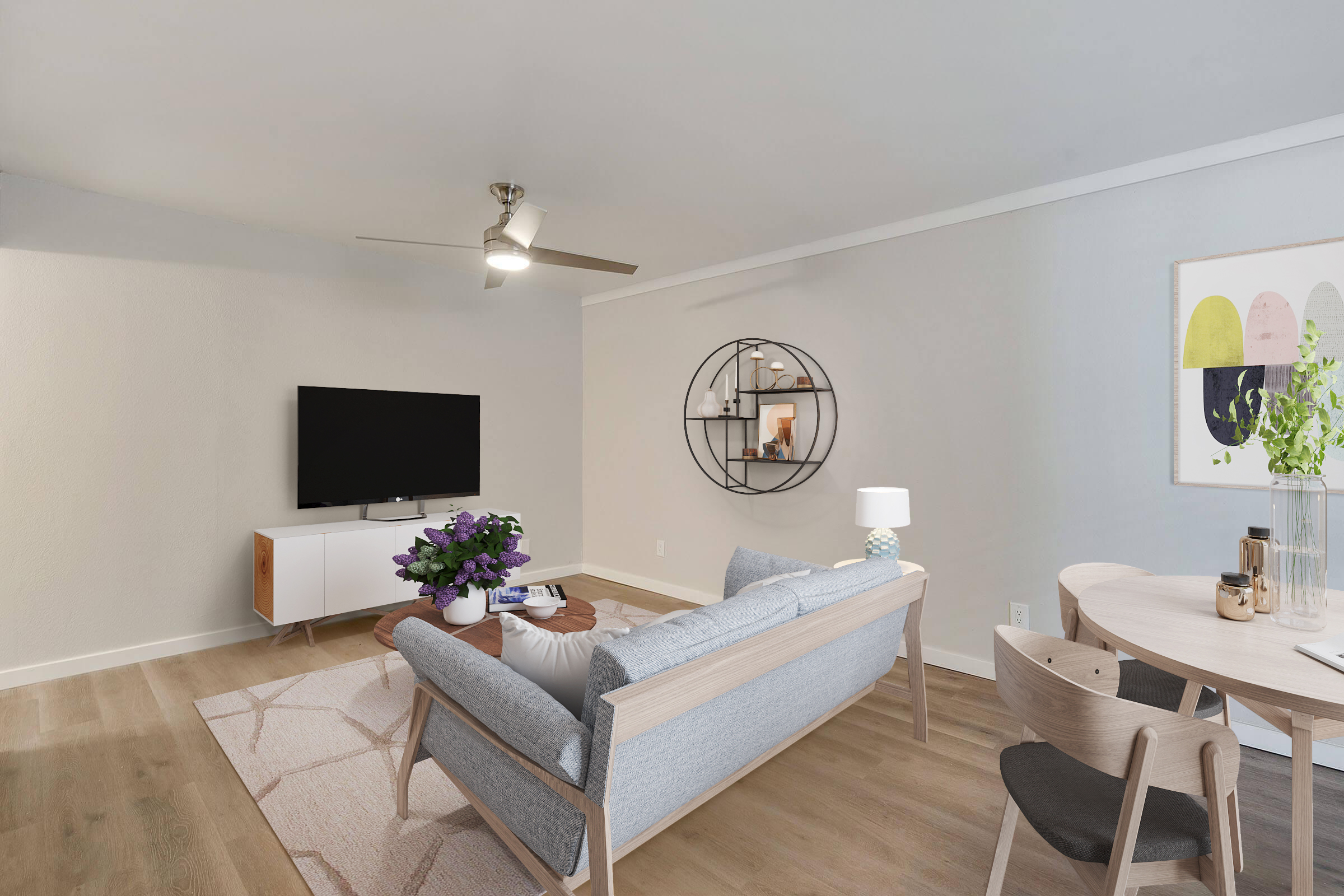 A modern living room featuring a light gray sofa with decorative pillows, a round coffee table, and a flat-screen TV mounted on the wall. There's a circular wall mirror and a small dining table with wooden chairs. Soft lighting and neutral colors create a bright, inviting atmosphere.