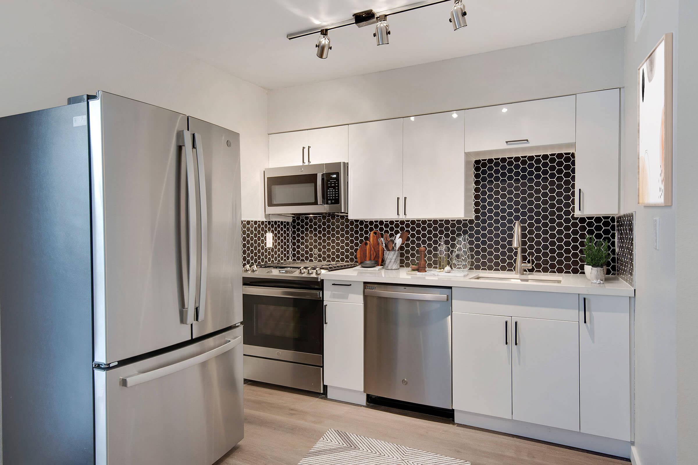 A modern kitchen featuring stainless steel appliances, including a refrigerator, oven, and microwave. The cabinetry is white with a sleek design, and a black hexagonal tile backsplash adds visual interest. The countertop is light-colored, complementing the overall contemporary aesthetic.