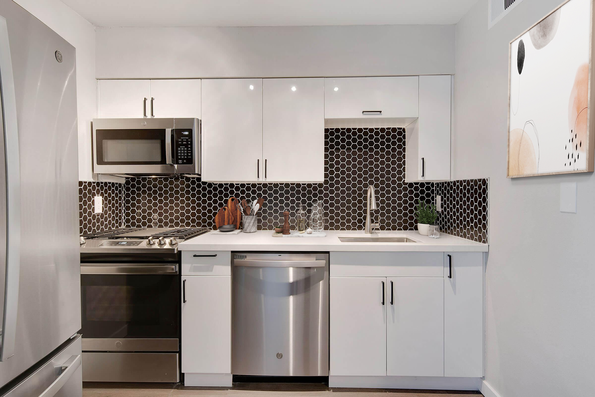 Modern kitchen featuring white cabinetry, stainless steel appliances, and a stylish hexagonal tile backsplash. The countertop is equipped with a sink, and there's a dishwasher beneath the counter. A framed abstract art piece hangs on the wall to the right. Well-organized and contemporary design.