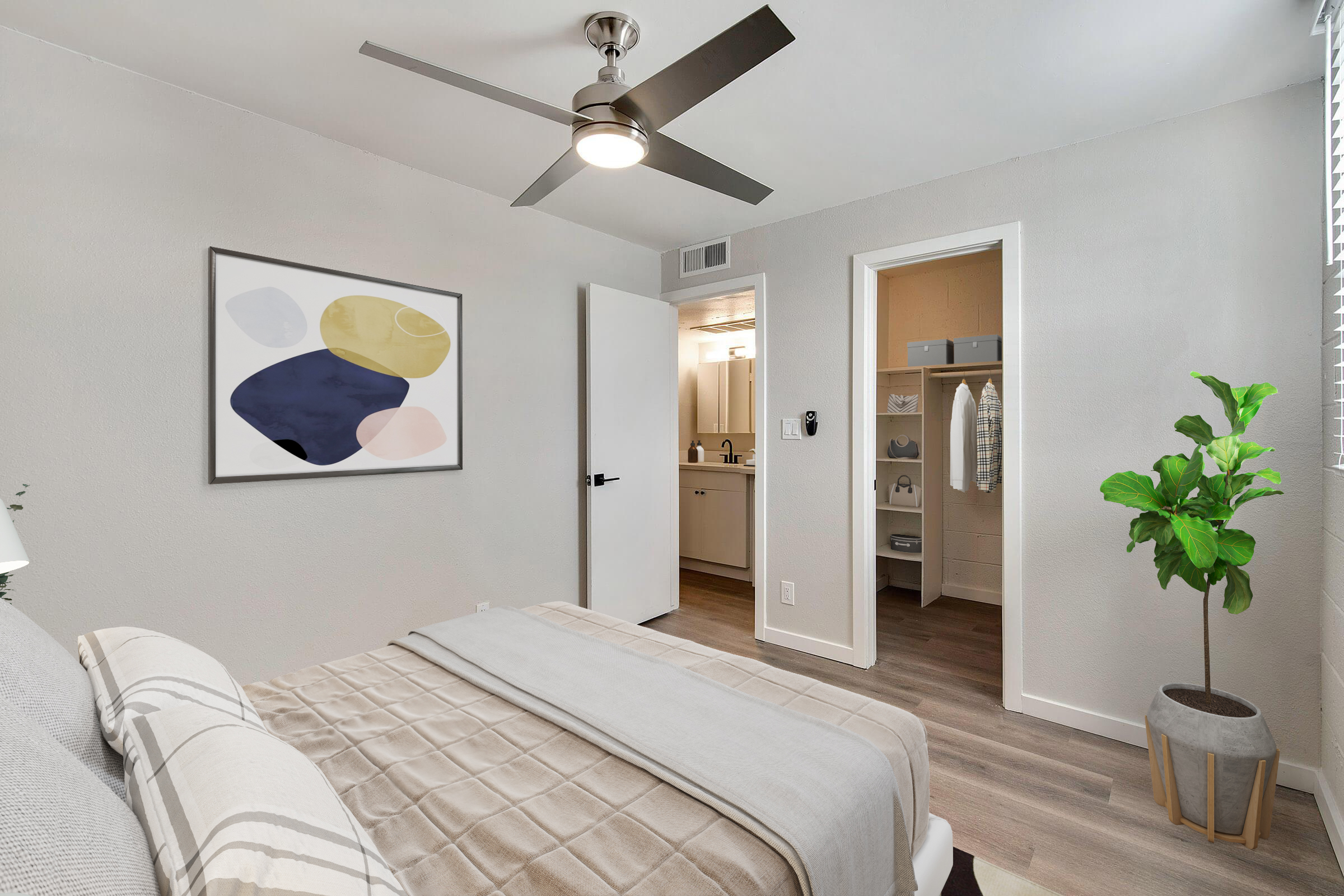 A modern bedroom featuring a neatly made bed with light bedding, a large abstract painting on the wall, a ceiling fan, and a potted plant in the corner. A door leads to an adjoining room, hinting at a connected bathroom or closet space, with neutral-colored walls and wood-like flooring.