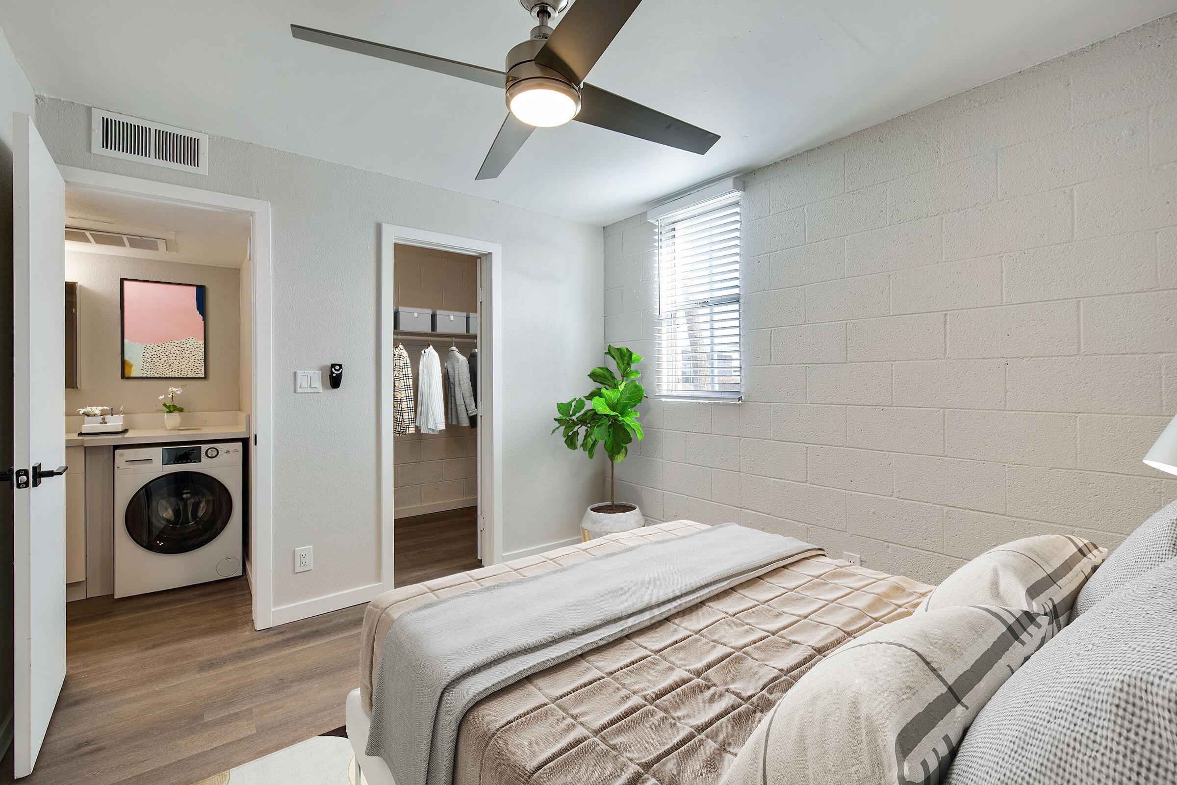 Cozy bedroom featuring a queen-sized bed with neutral bedding, a ceiling fan, and a window providing natural light. A small plant adds a touch of greenery. The room connects to a hallway with a washer and dryer visible, along with an adjacent room that has a closet with clothes hanging.