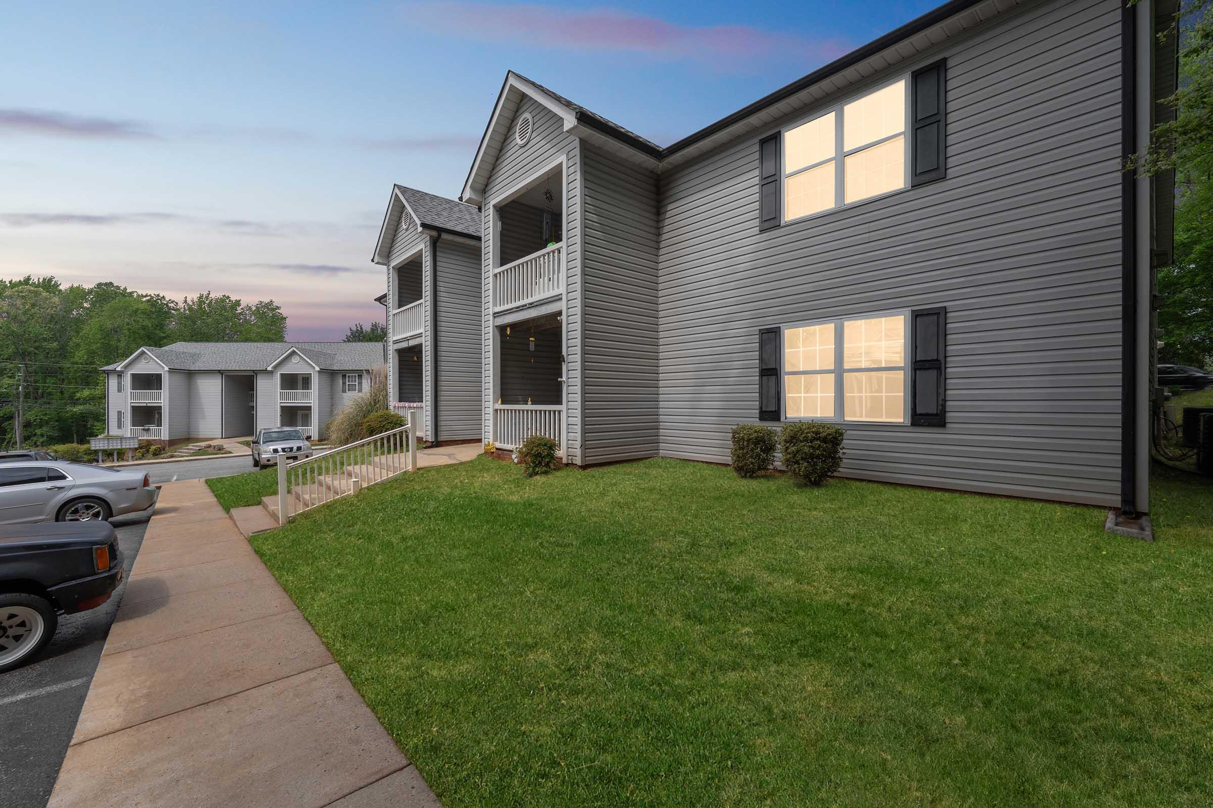 A two-story apartment building with gray siding. The building features a porch with white railings and several windows, some lit up. Lush green grass surrounds the building, and there are a few parked cars nearby. The sky is visible in the background, hinting at twilight.