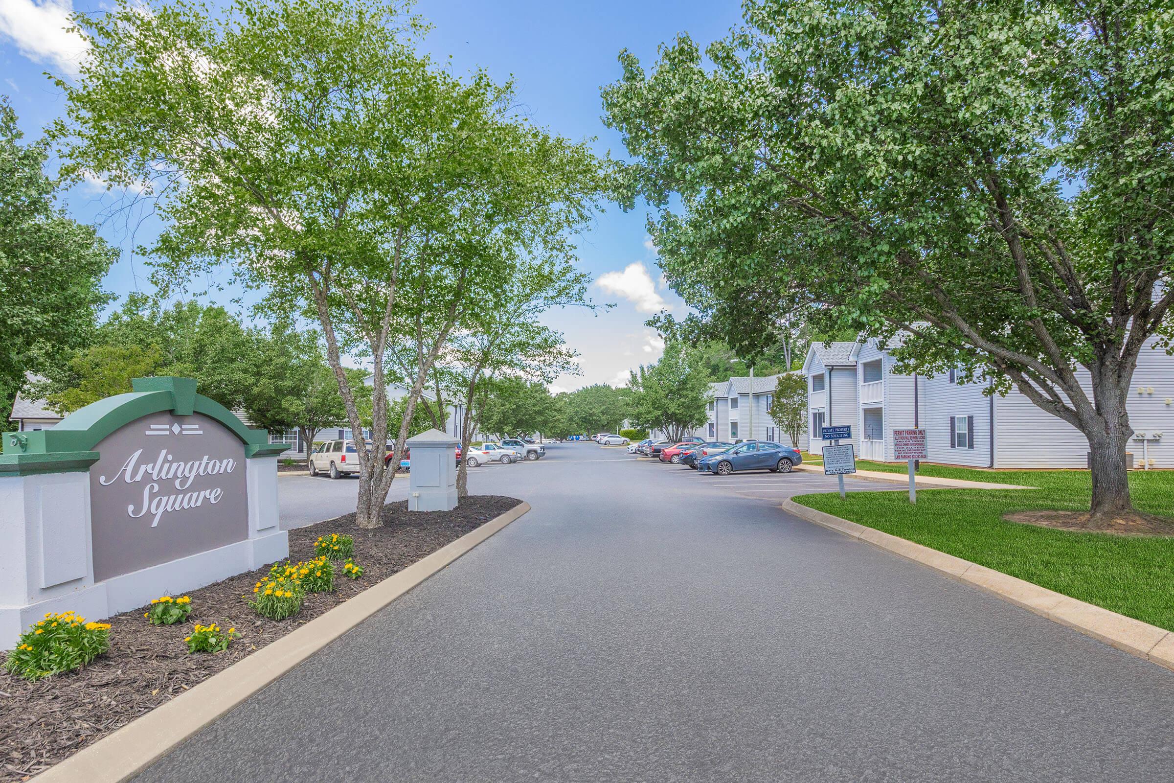 A sunny view of Arlington Square, featuring a paved road lined with greenery and trees. The community sign is visible on the left, and residential buildings can be seen along the road on the right. Parking areas are also present, creating a welcoming atmosphere.