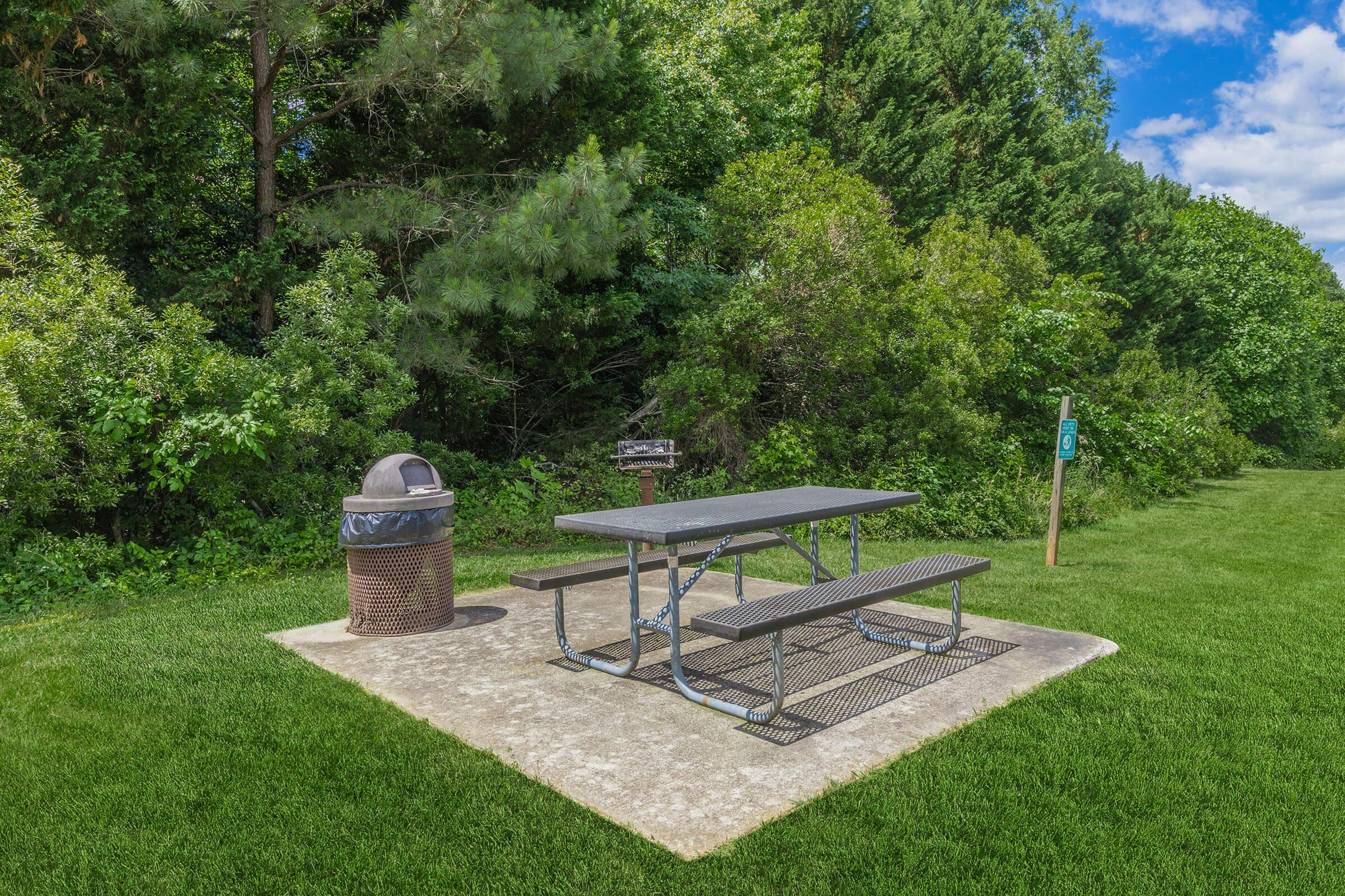 A picnic table with metal benches is situated on a concrete pad, surrounded by lush green trees and grass. Next to the table, there is a trash can, and a sign visible in the background. The scene is bright and sunny, suggesting a pleasant outdoor area for gatherings or relaxation.
