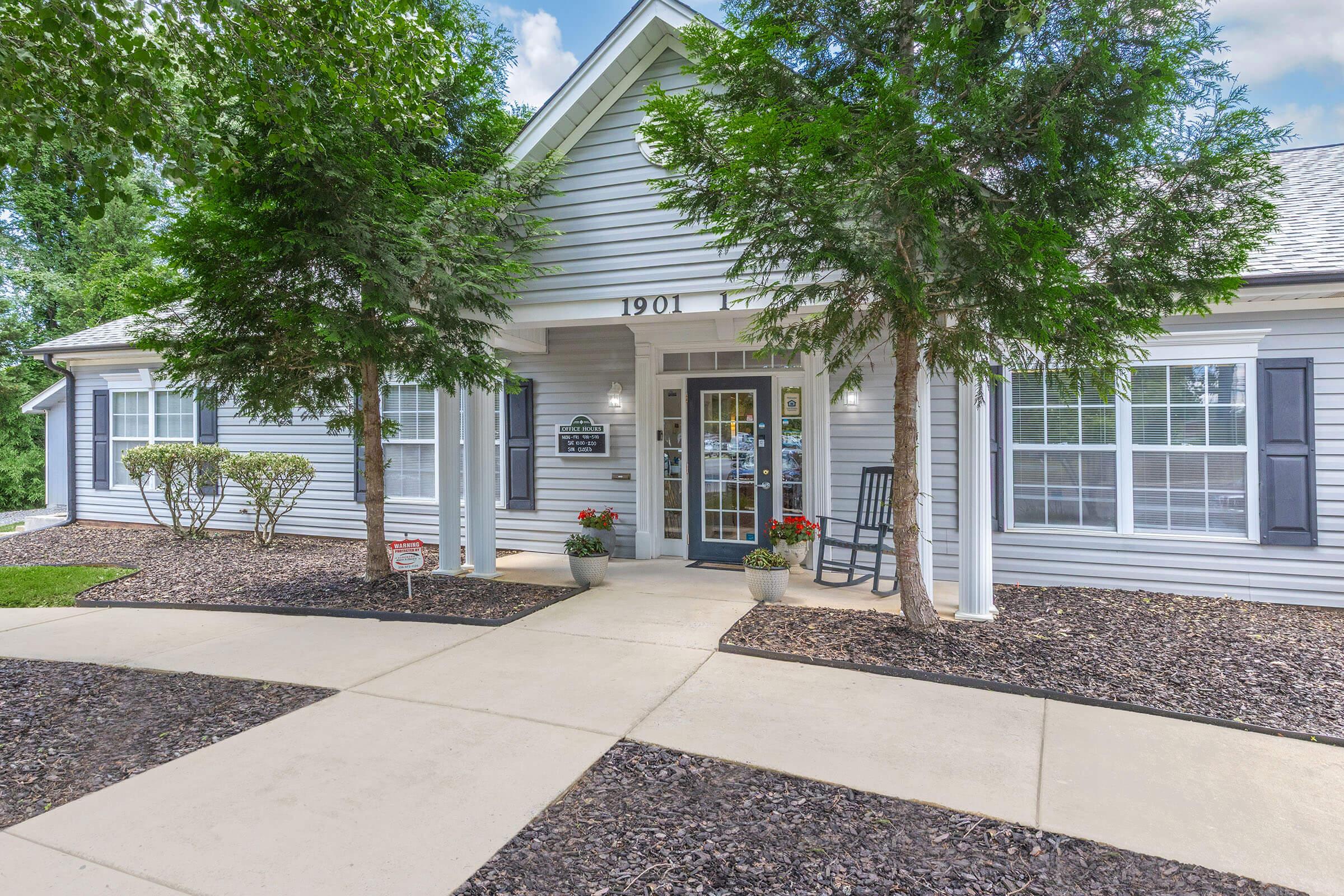 A well-maintained exterior of a single-story house featuring a light gray siding, white trim, and a welcoming entrance. The path is bordered by mulched areas and shrubs, with potted plants by the front door. A sign indicates the house number "1901," surrounded by green trees and a blue sky.