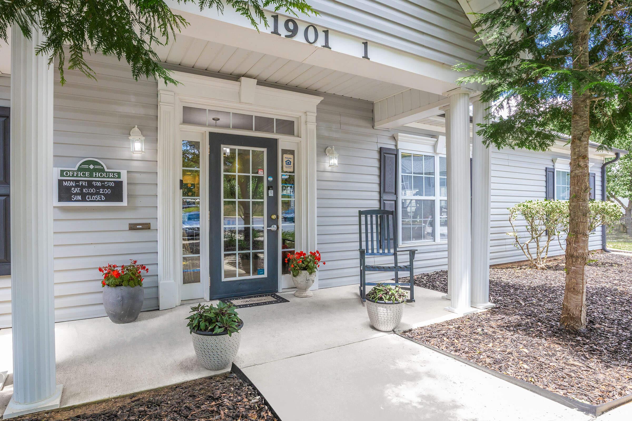 Exterior view of a welcoming office entrance with a front porch. The door is flanked by tall potted plants and a small bench. A sign displaying office hours is visible next to the door. The building is light gray with white trim, and there are trees nearby, adding a touch of greenery to the setting.