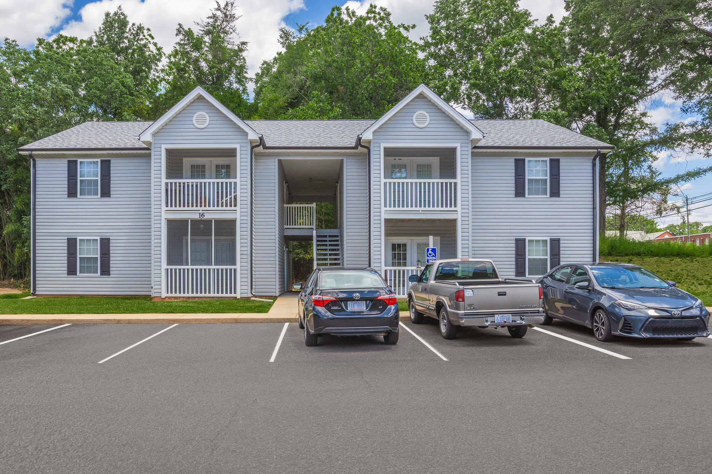 Two-story apartment building with grey siding and a pitched roof, featuring balconies on the second floor. A parking lot in front holds three cars, including a grey pickup truck and two sedans. Lush green trees surround the building, under a blue sky with scattered clouds.