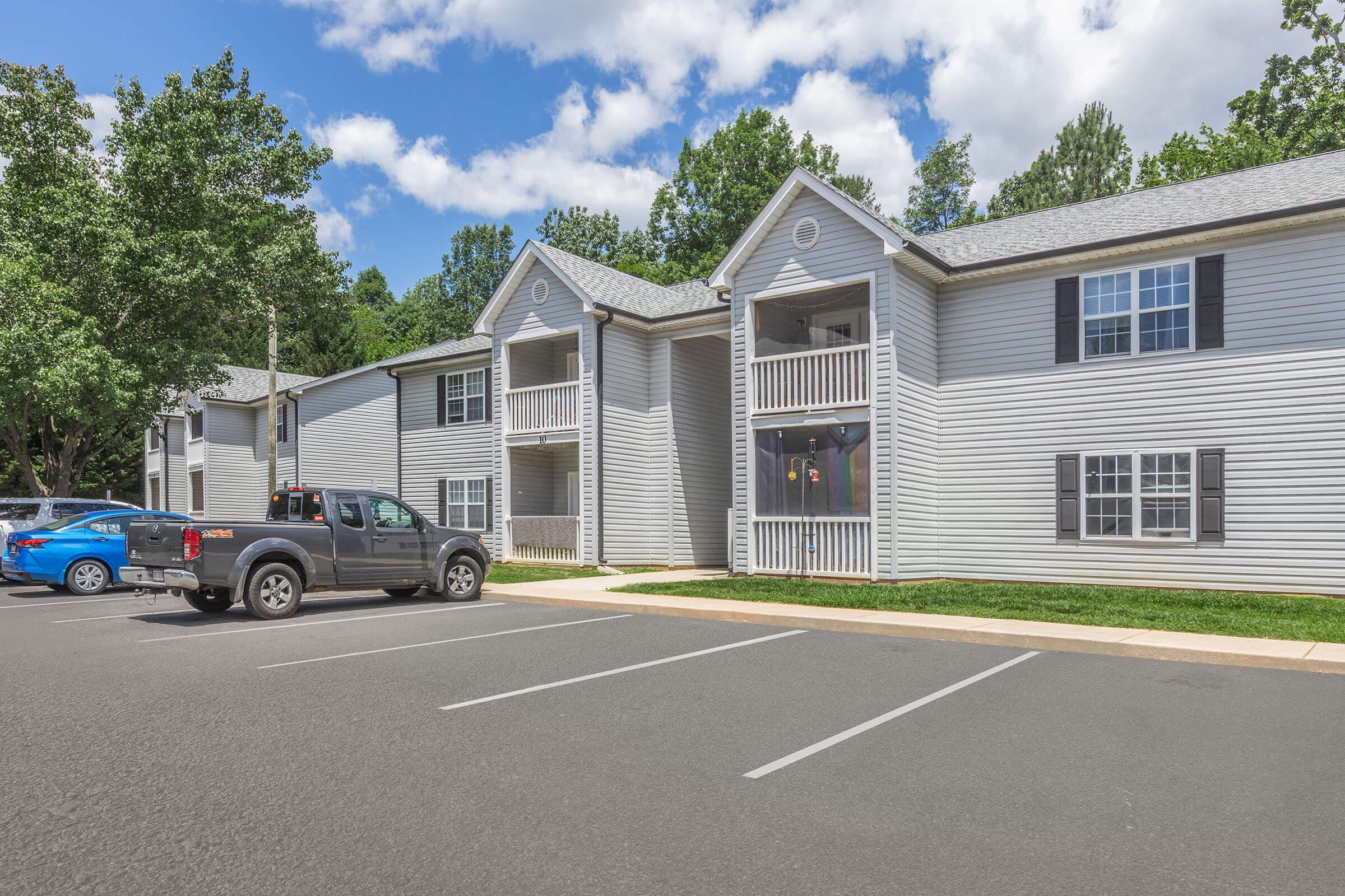 Two-story residential apartment building with gray siding, featuring balconies on the second floor. The foreground shows a parking area with a blue car and a pickup truck, surrounded by well-maintained greenery and trees under a partly cloudy sky.
