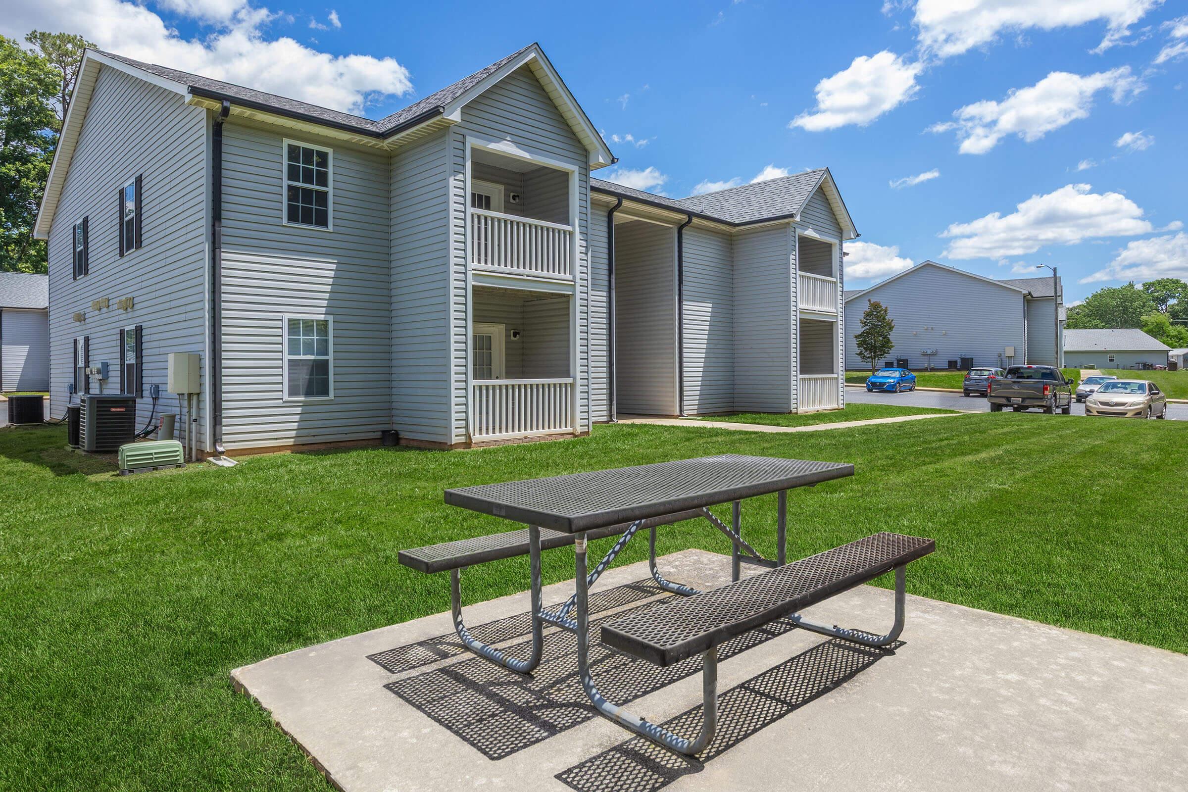 A bright, sunny day view of an apartment complex with two buildings. In the foreground, there is a picnic table on a concrete slab surrounded by green grass. The buildings feature multiple balconies and are set against a backdrop of blue skies with scattered clouds.